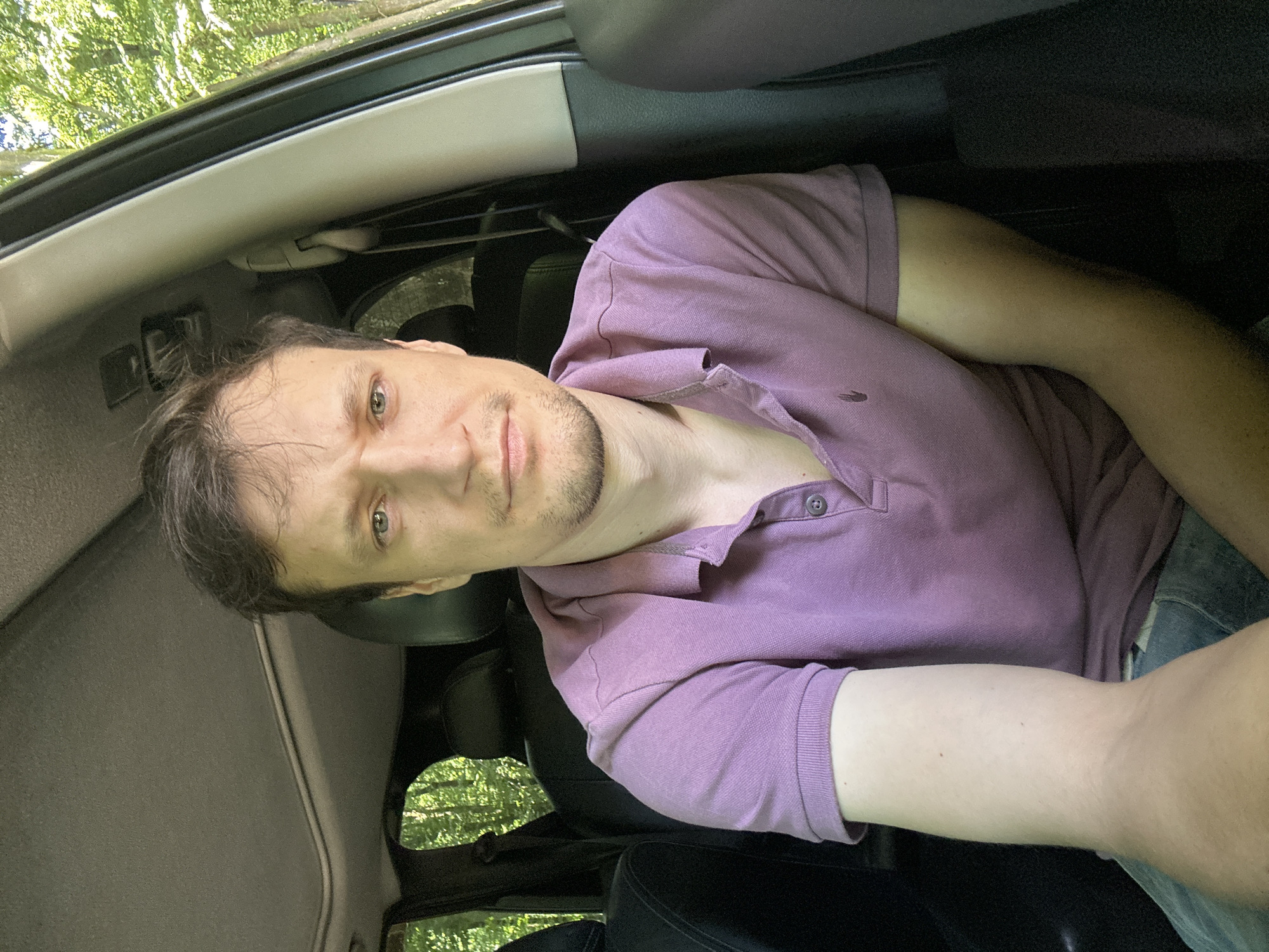 A medium shot shows a man sitting in the front passenger seat of a car. He has light brown hair, fair skin, and light green eyes. He is wearing a short-sleeved, lavender polo shirt with a small logo on the left side of the chest. The car's interior is visible, including the dark headliner, a sunroof, and the backs of the seats. Through the windows, a wooded area with lush green foliage is visible, suggesting the car is in a rural or natural setting. The man's expression is neutral, and he is looking directly at the camera.