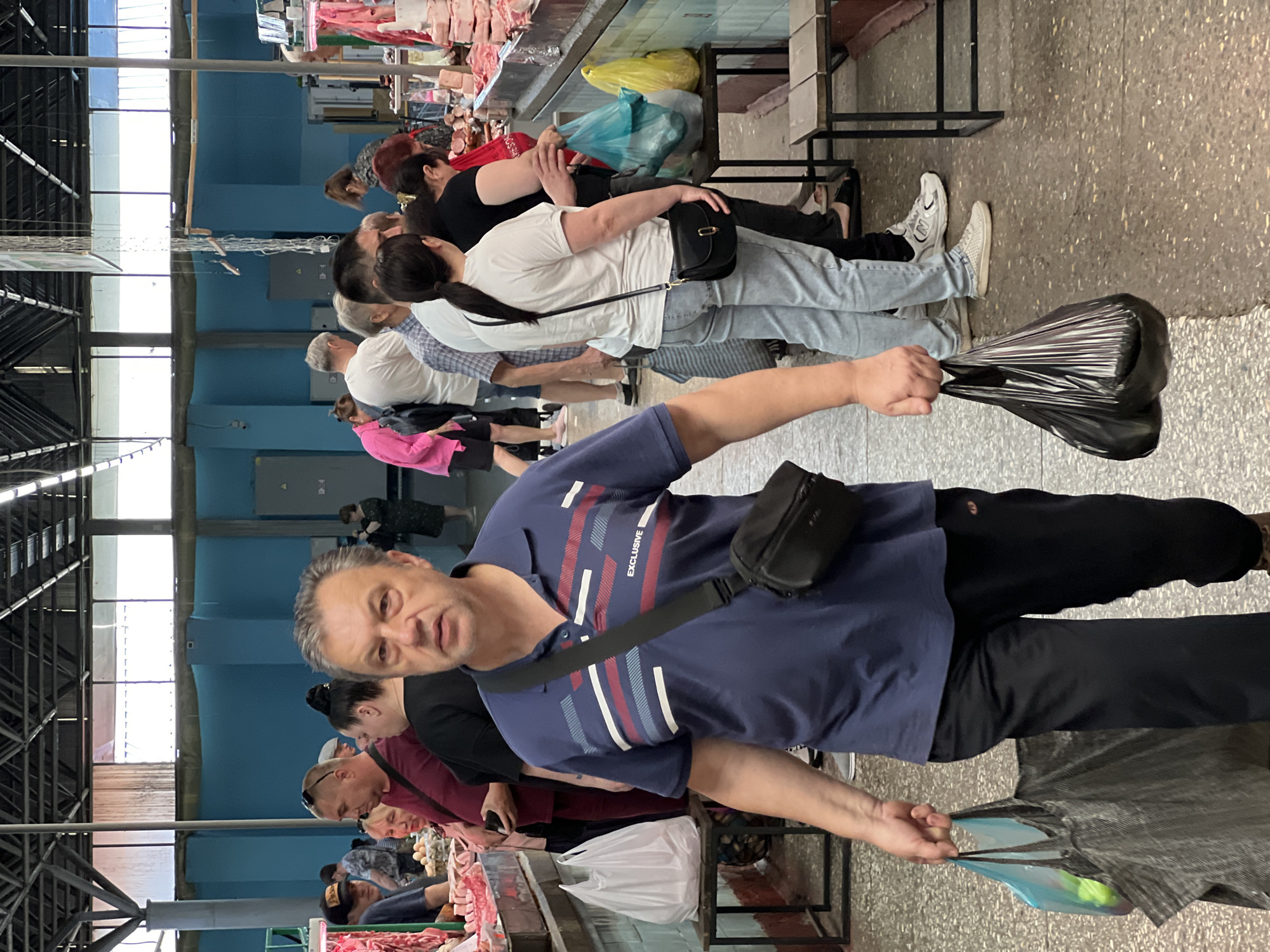 This image depicts an indoor market in Krasnodar, Russia, characterized by its industrial ceiling structure and light blue walls. The scene is moderately crowded with shoppers navigating the aisles between various stalls.
In the foreground, a middle-aged man with short, grey hair and a neutral expression walks directly towards the viewer. He is wearing a dark blue polo shirt with horizontal white, red, and light blue stripes across the chest, and the word "EXCLUSIVE" printed in white on the right side of his chest. A black cross-body bag hangs over his right shoulder. He wears dark athletic trousers with a small, indistinct logo on the right thigh. In his left hand, he carries two shopping bags: a translucent blue plastic bag and a larger, dark grey/black reusable fabric bag.
To the man's left, a market stall displays various cuts of raw meat on a metal counter. A white plastic shopping bag rests on a small wooden stool next to this stall. Further back on the left, other shoppers are visible, including a man with sunglasses perched on his head and a woman with blonde hair. Trays of eggs are also partially visible on a counter in the background.
To the man's right, another market aisle features more stalls with raw and processed meat products. Several shoppers are gathered at these stalls, including a woman seen from behind, wearing a white t-shirt, light blue jeans, and a black cross-body bag. Next to her, a woman in a black top is reaching towards the display. On a wooden stool near these stalls, a translucent blue plastic bag and a yellow plastic bag are placed. In the distant background, other people are seen walking and standing.
The market features a grey concrete floor, light blue painted walls, and an exposed metal truss ceiling with large, high-set windows that provide natural light. Grey electrical panels are mounted on the walls in the background. The overall impression is that of a bustling, functional market environment.