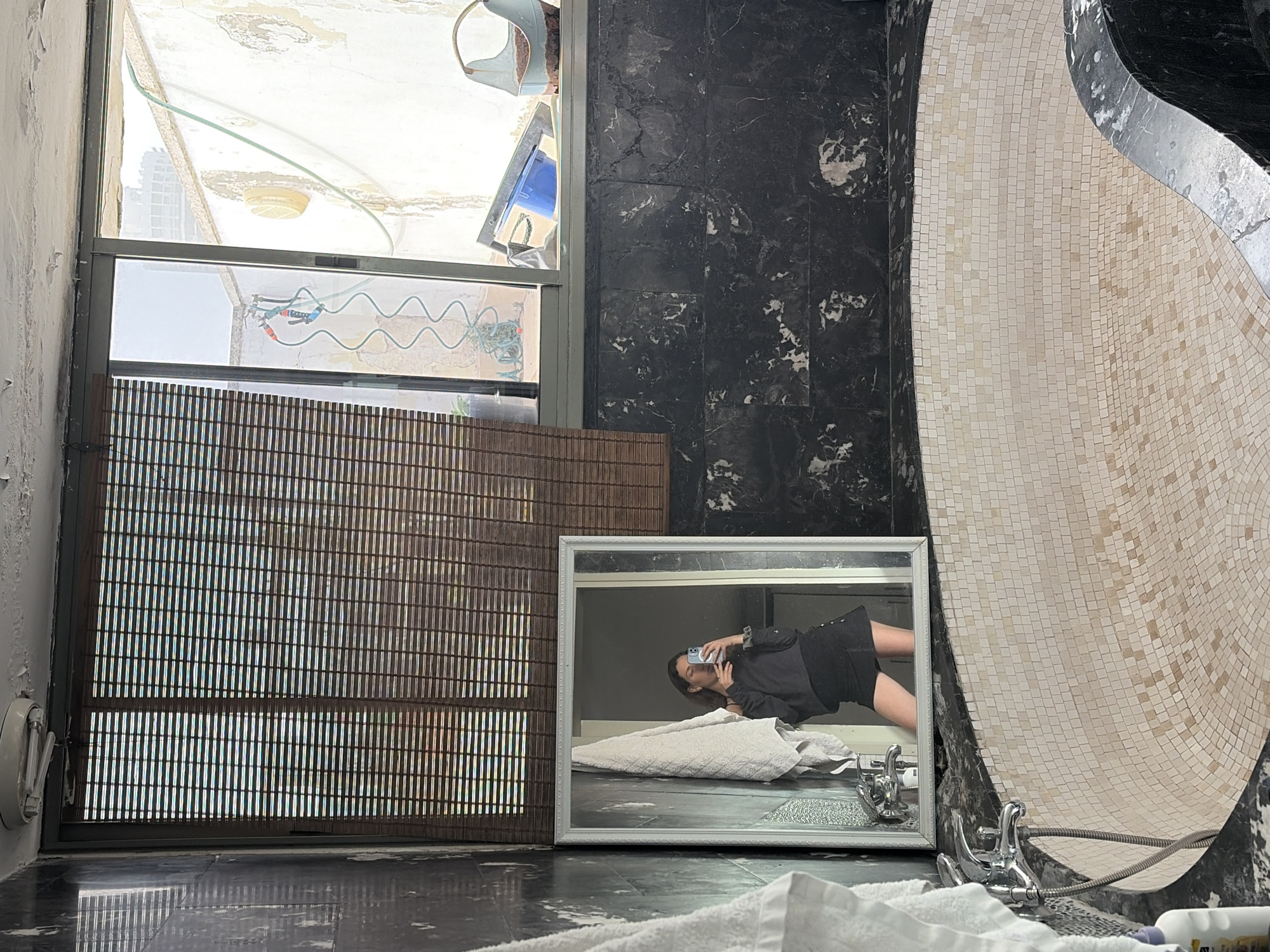 The image presents a scene inside an apparent bathroom or washroom area, likely under renovation or construction, suggested by the unfinished or worn walls and visible building materials outside the window.
Through a window with a brown slatted blind, an exterior view is discernible, displaying features of the city of Tel-Aviv, Israel. The view includes urban architecture and the sky above the city. Plumbing and other construction materials are visible outside the window.
A woman is reflected in a mirror, which is resting on the floor, taking a photo of herself with a mobile phone. She appears to be lying on a towel or padding, wearing a dark top and shorts.
The bathroom itself is characterized by contrasting textures; a section of wall is covered with dark, marbled tiles, adjacent to a decorative element constructed with light-colored mosaic tiles. Chrome bathroom fixtures, including taps and a shower head, are present near the tiled wall.
The floor is dark and reflective, suggesting it may be wet or recently cleaned. A white towel is visible in the foreground, partially obscuring the lower part of the frame.