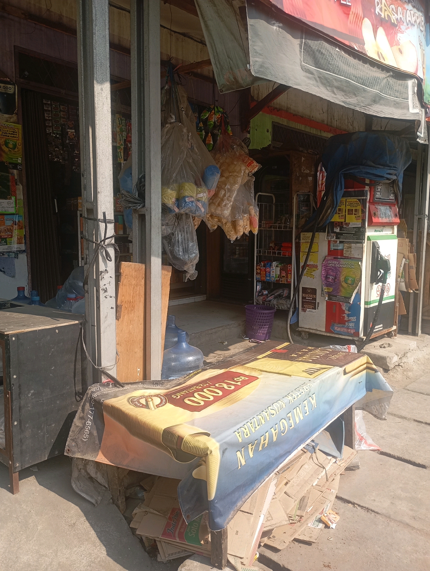 The image depicts an outdoor scene in Babelan, Indonesia, likely in front of a small shop or warung. A table covered with a large banner displaying text and images is positioned in the foreground. Underneath the table, stacks of cardboard boxes and other debris are visible. To the left of the table is a black cabinet or box. Behind the table, a shop entrance is partially visible, with shelves stocked with various items and plastic bags of snacks hanging. A purple basket is placed on the ground. The environment appears to be a sunny day, with shadows cast on the concrete ground. The overall scene suggests a commercial area with goods being sold or stored.