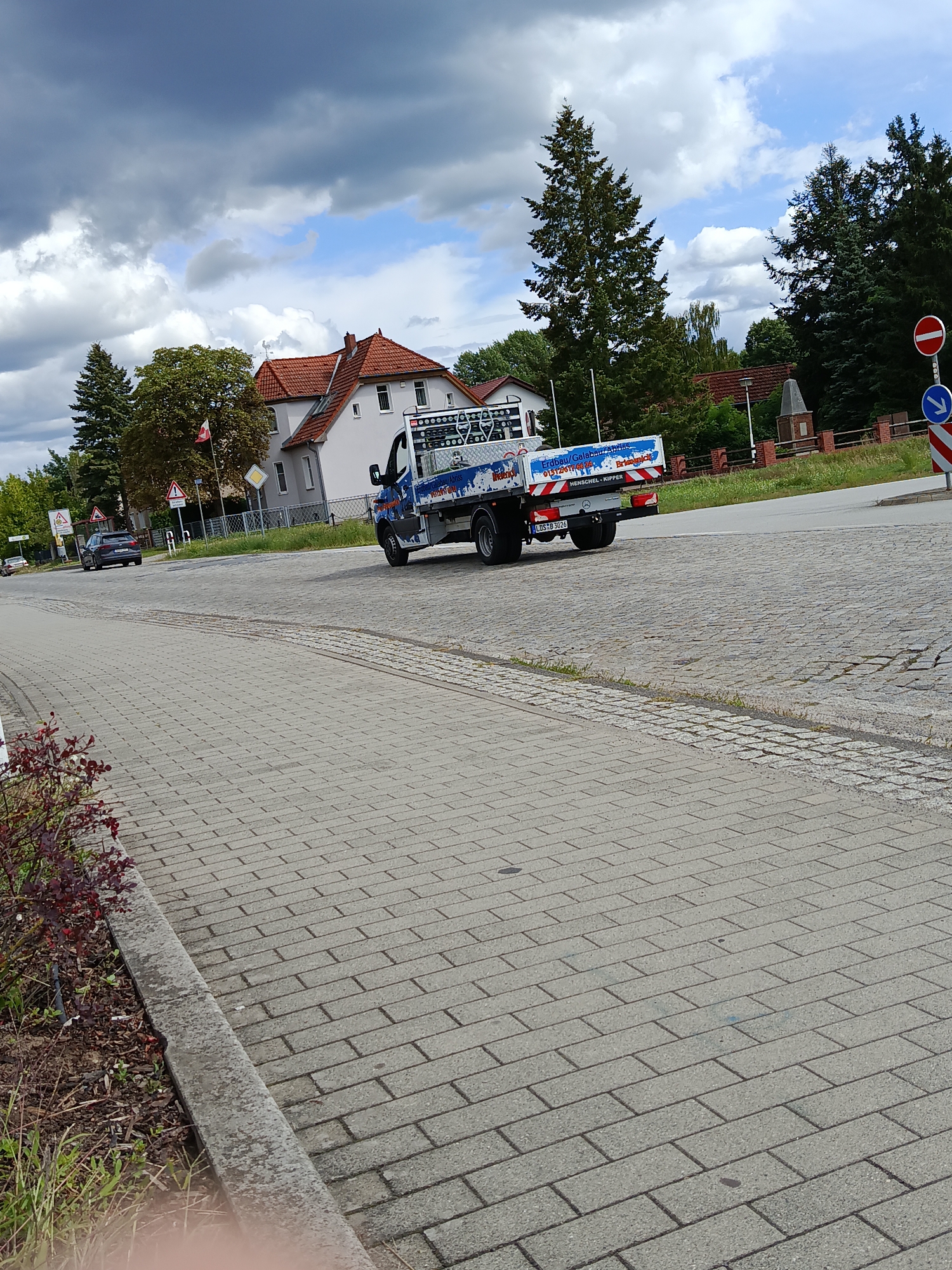 The image shows a street scene in Groß Köris, Germany, under a cloudy sky. A blue and white truck with advertisements on its side is driving on the road. The truck's license plate reads "LDS B 3026". In the background, there are houses with red roofs, trees, and street signs. A grey car is parked on the side of the road. The foreground shows a paved sidewalk with some plants and bushes on the left. The overall impression is that of a typical day in a suburban area.