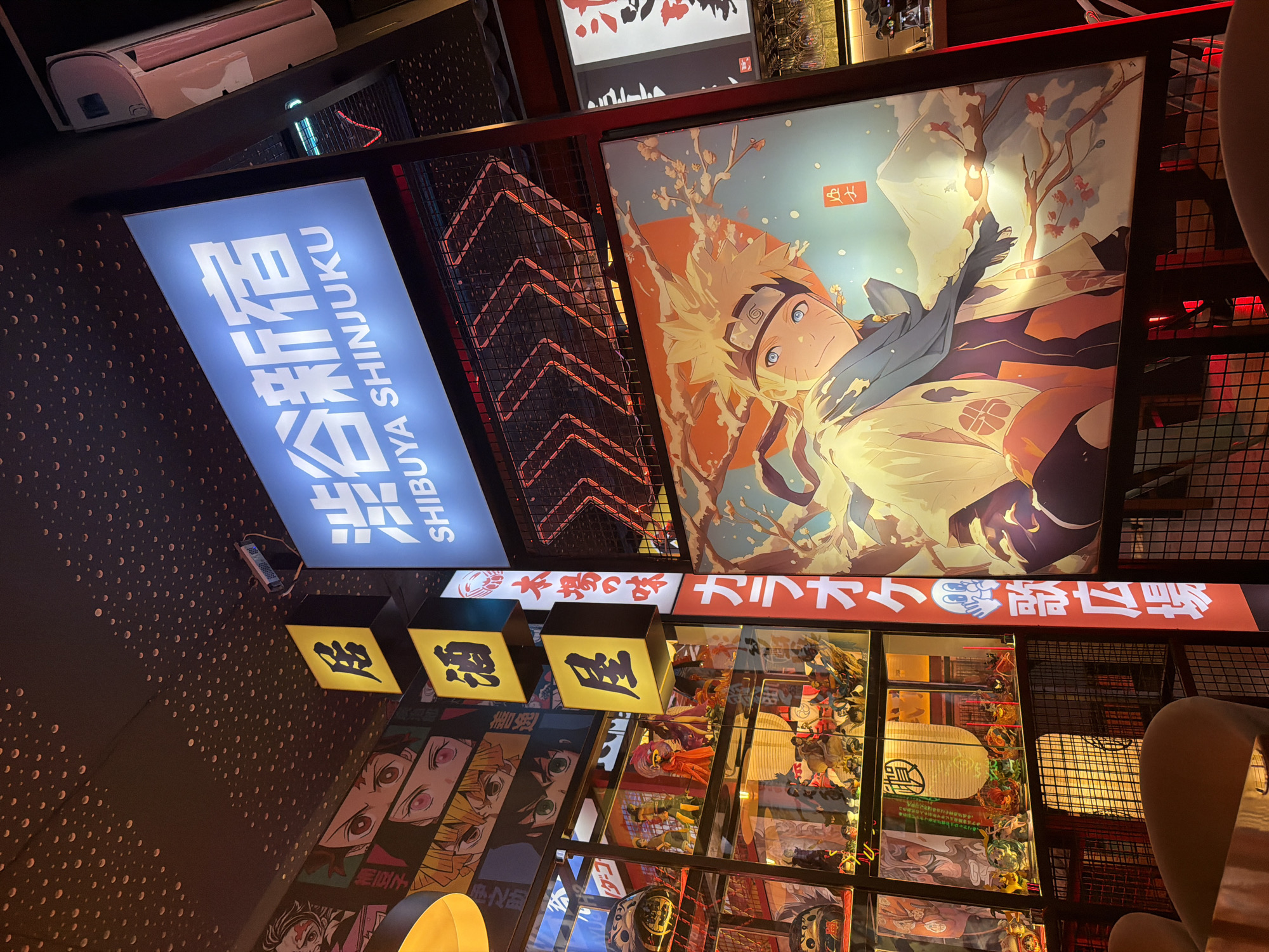 The image depicts the interior of an anime-themed restaurant or bar, likely located in Lisbon, Portugal. The scene is vibrantly decorated with Japanese cultural elements and anime imagery. A prominent blue illuminated sign in the upper right corner displays Japanese characters and the text "SHIBUYA SHINJUKU". Below this, a large poster features an anime character with blonde spiky hair and blue eyes, possibly Naruto, with a snowy mountain and cherry blossoms in the background.
To the left, stacked yellow square signs with black Japanese characters hang vertically, indicating "居" (kyo or i - dwelling/restaurant) and "酒" (shu - alcohol/sake). Further to the left, a wall is adorned with a collage of anime character eyes and faces, featuring styles reminiscent of popular series like Demon Slayer.
A glass display case filled with various anime figurines and decorative items, including traditional Japanese lanterns, is visible on the left side of the image. Vertical red signs with white Japanese characters, likely advertisements or signage for the establishment, run along the right edge of the display case. The ceiling is dark and textured with small, circular, raised patterns. The lighting is a mix of neon and ambient light, casting a warm and colorful glow on the displays and creating a lively atmosphere. Chairs and tables, partially visible in the foreground, suggest a seating area for patrons.