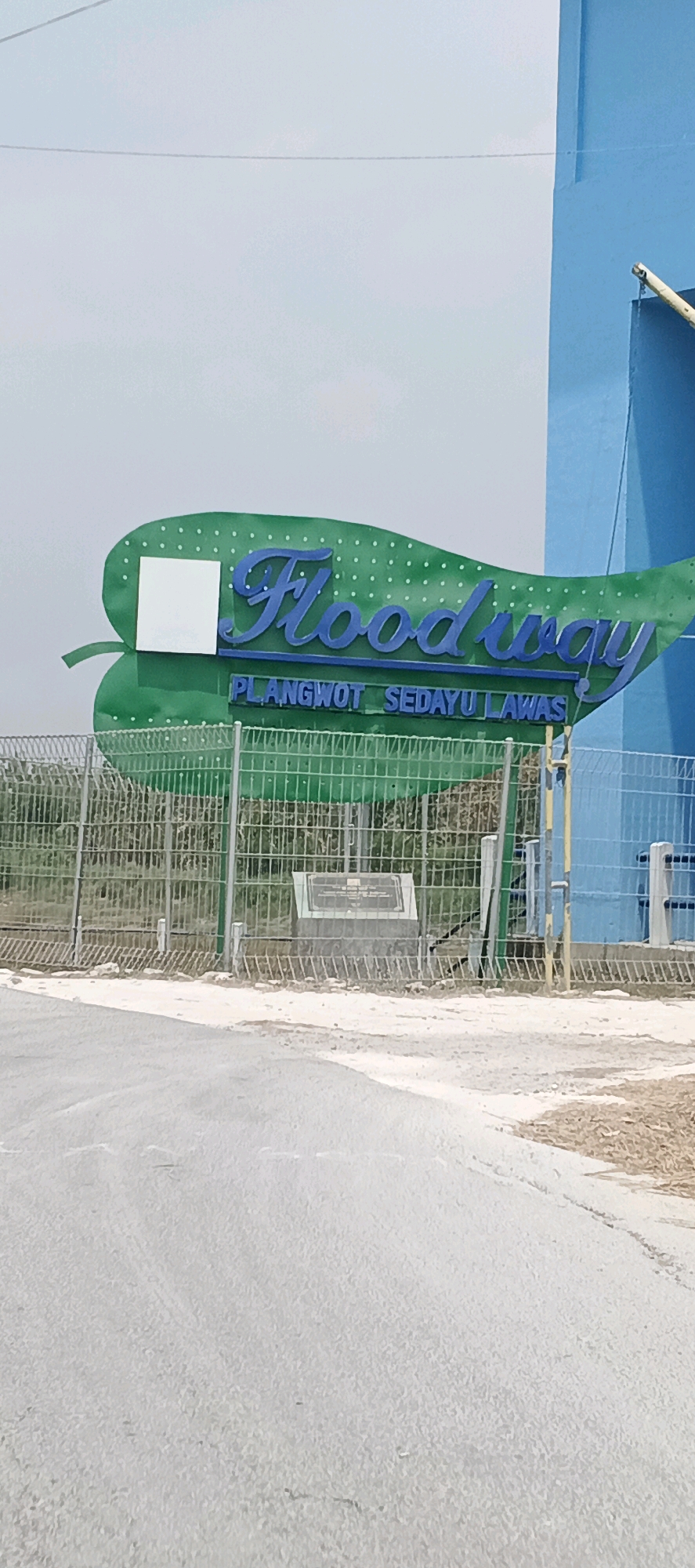 The image shows a large green sign shaped like a leaf with the words "Floodway" in blue cursive lettering. Below "Floodway", smaller blue text reads "PLANGWOT SEDAYU LAWAS". A white square is affixed to the left side of the leaf sign. Behind the sign, a chain-link fence is visible, and further back, a light blue building stands. A concrete plaque with text is mounted on a stone base behind the fence. The scene appears to be outdoors, with a dirt or gravel path in the foreground and a pale sky in the background. The location is identified as Parengan, Indonesia.