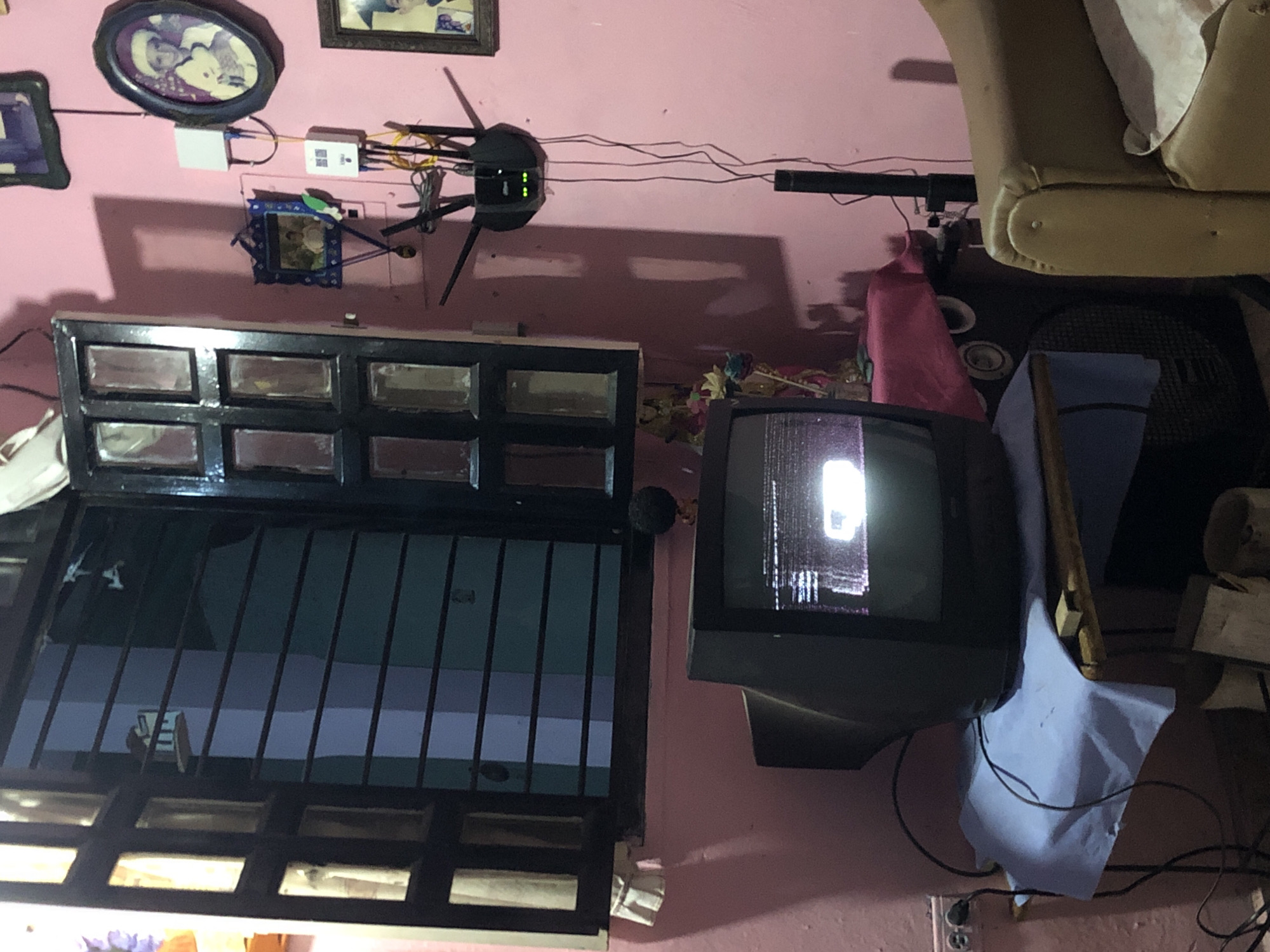 The image shows an indoor scene with a pink wall as the backdrop. Several framed pictures are hanging on the wall, along with a router and some network equipment. Below the decorations, there's a black window with multiple panes and bars. To the right of the window, there's a television set with a lit screen. Beside the TV, there's an electric fan, partly covered by a light blue cloth. The top right corner of the image features a part of a beige armchair. Cables and wires are visible near the floor. The city of Altagracia, Venezuela, is not discernible from the image.