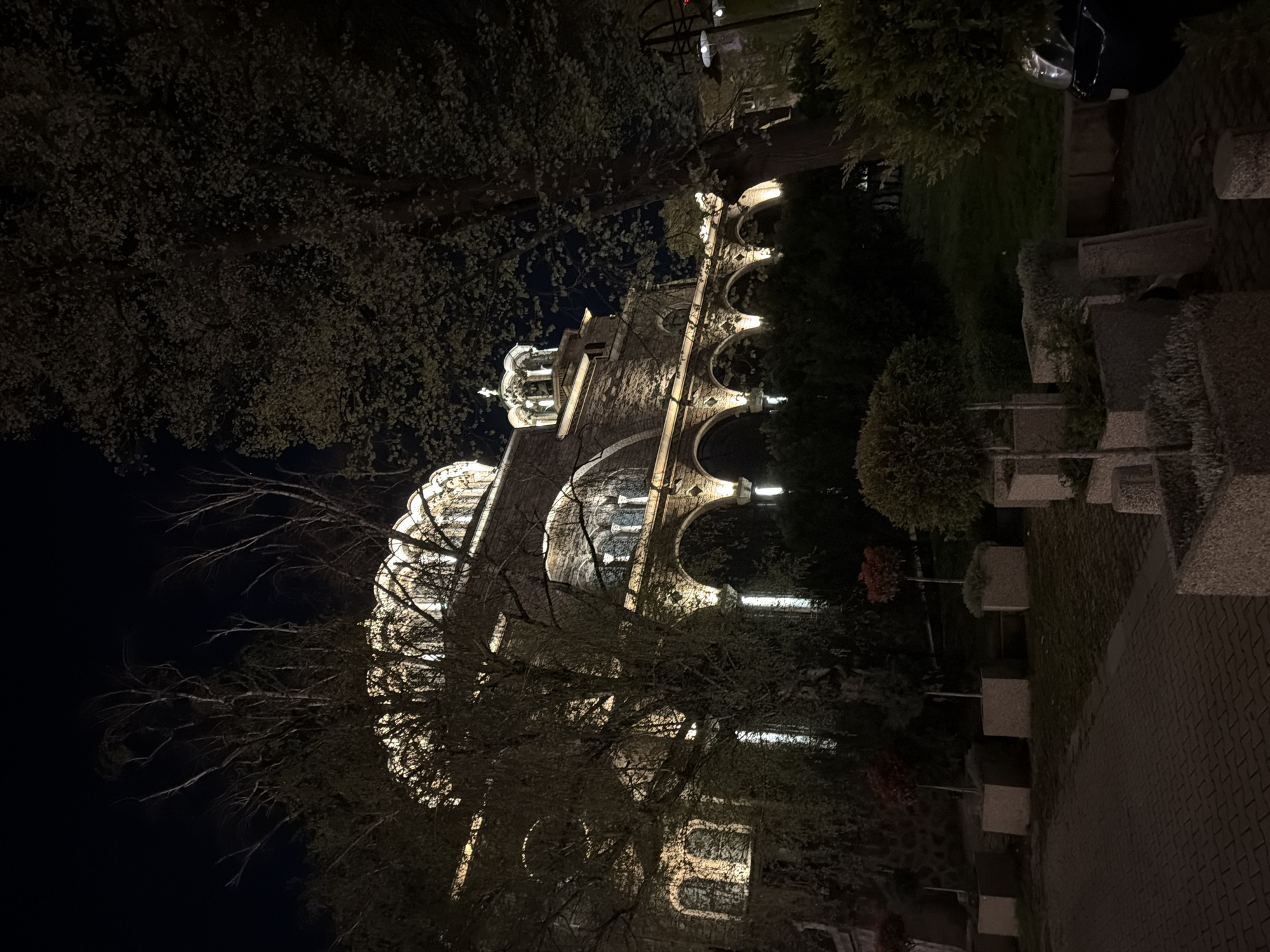The image shows a building with arched windows and a tower, illuminated at night. The building is surrounded by trees, and a paved area is visible in the foreground with square planters. The location is Unknown, Unknown.