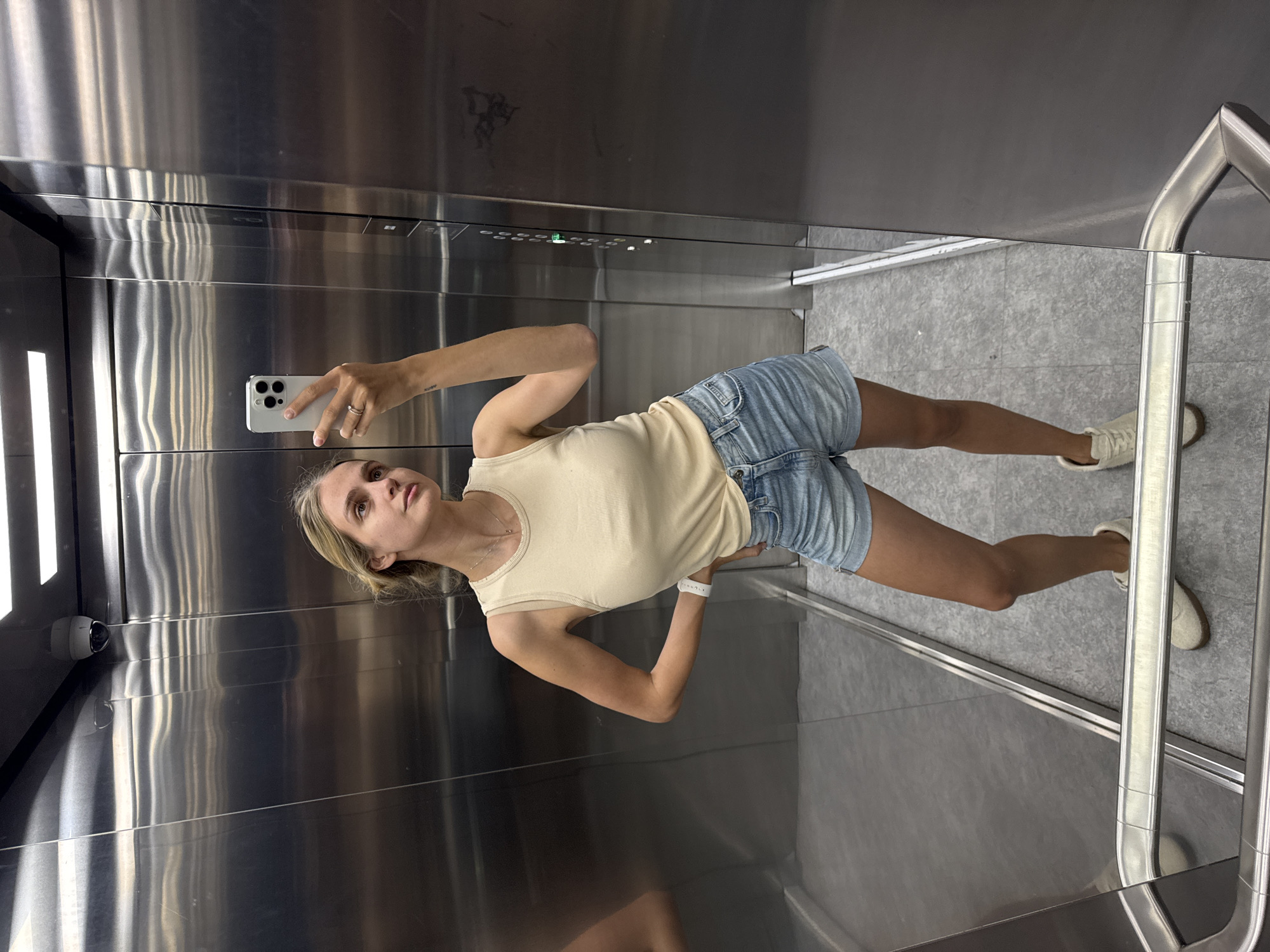 The image shows a woman taking a selfie in an elevator. She is wearing a light-colored tank top and denim shorts, and white sneakers. The elevator has stainless steel walls and a gray tiled floor. A handrail is also visible. A security camera is installed near the door.