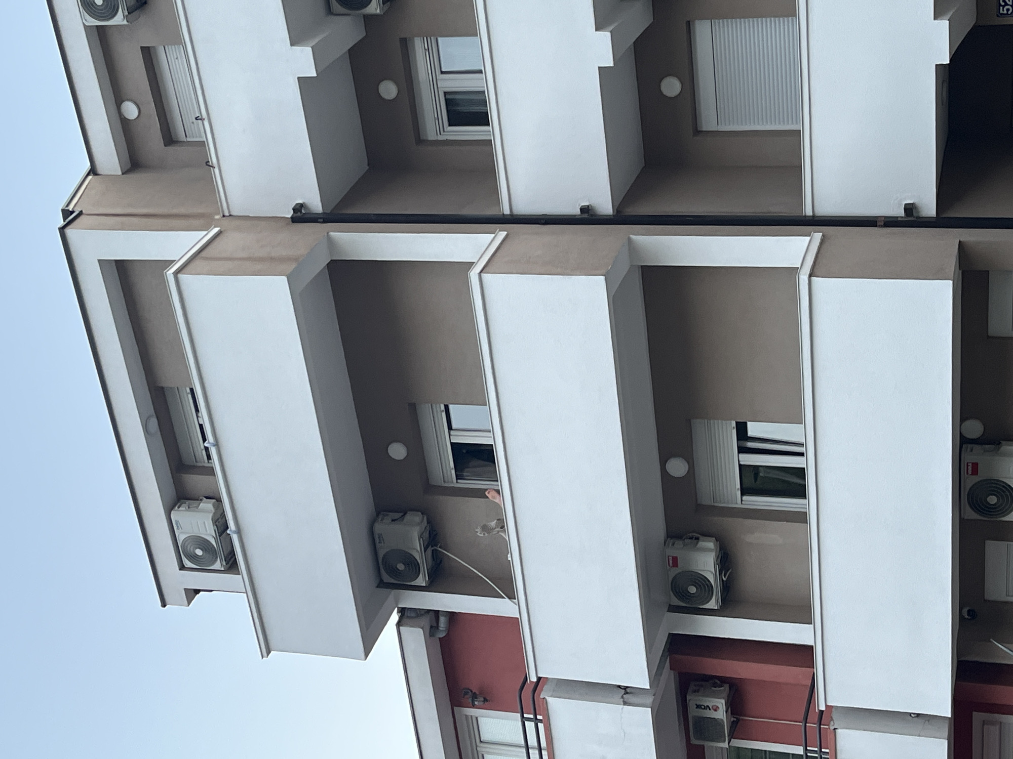 The image captures a section of a multi-story apartment building with a clear sky in the background, likely taken from a street level perspective. The building features balconies with white railings and brown walls. Several outdoor air conditioning units are mounted on the exterior walls. On one of the balconies, a person's legs and feet are visible, leaning over the railing. A cat is also present on the same balcony, near the person. The overall impression is a residential scene in a European city, possibly Budva, Montenegro, given the context.