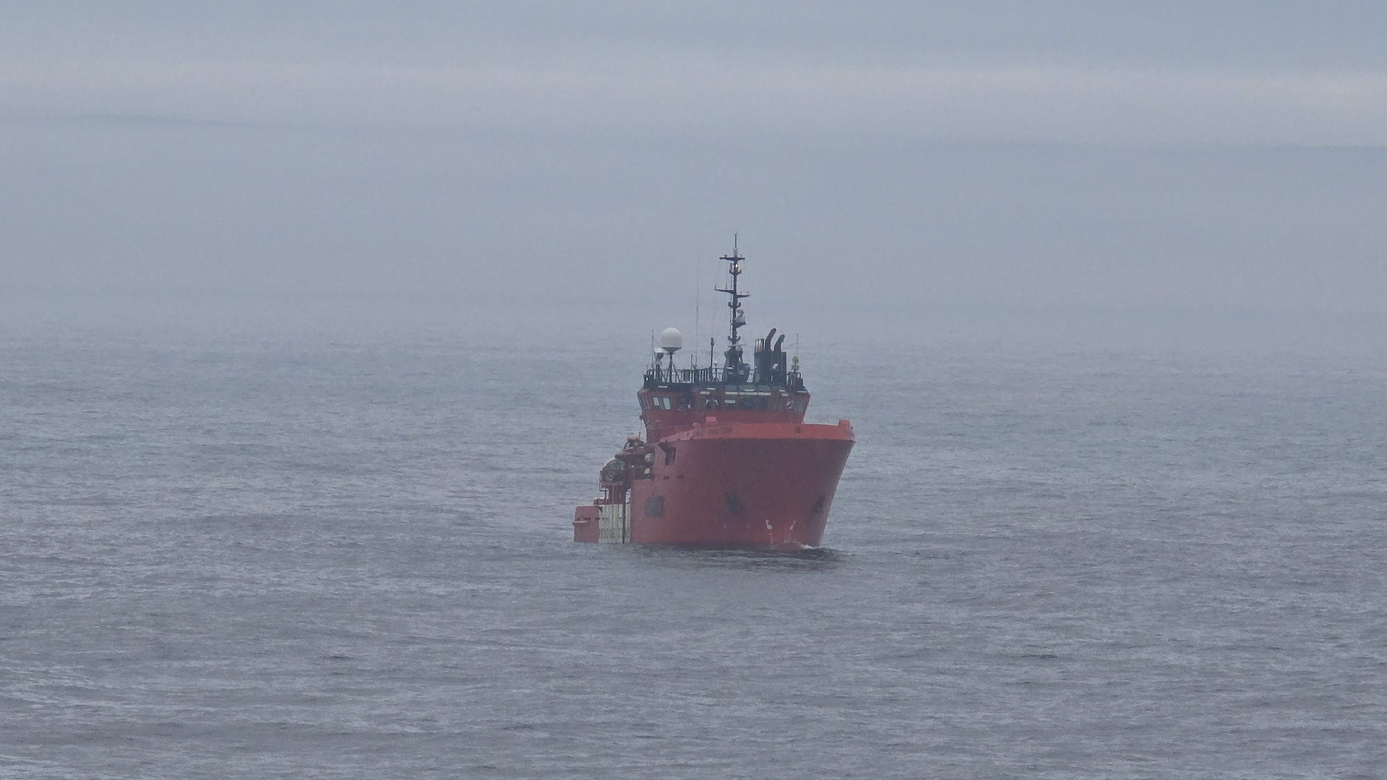 A red vessel is on the water under a gray sky. The vessel has a white dome radar antenna and multiple smaller antennas on top of the pilot house. The vessel is in a body of water that is gray. The sky above is gray. The location is Unknown, Unknown.