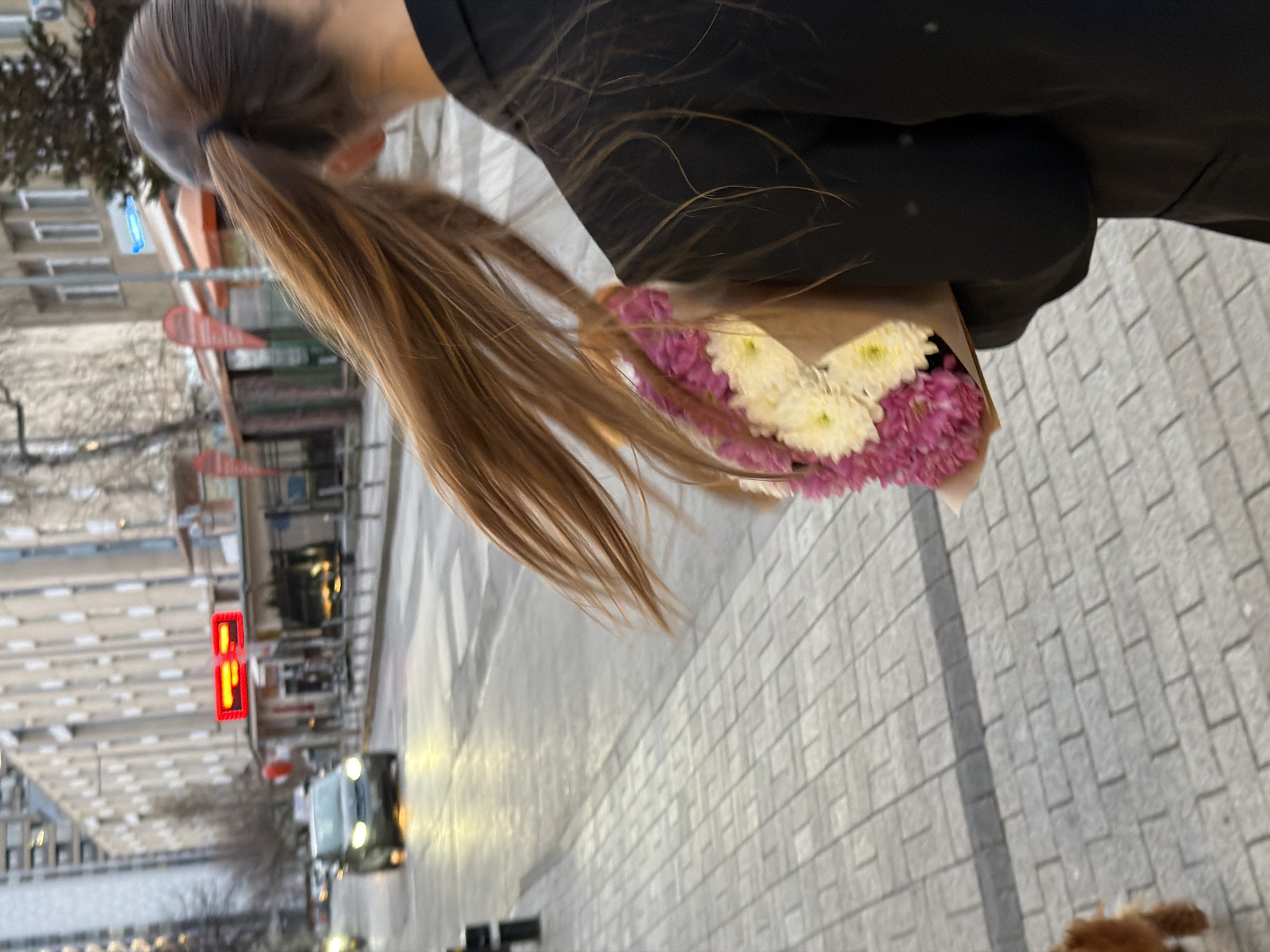 A woman with brown hair in a ponytail holds a bouquet of white and pink flowers, walking on a light gray brick sidewalk. The background shows a city street with buildings, a crosswalk, and a car waiting at a red traffic light. Based on architectural styles and street signs, the location is identified as being in Sofia, Bulgaria.