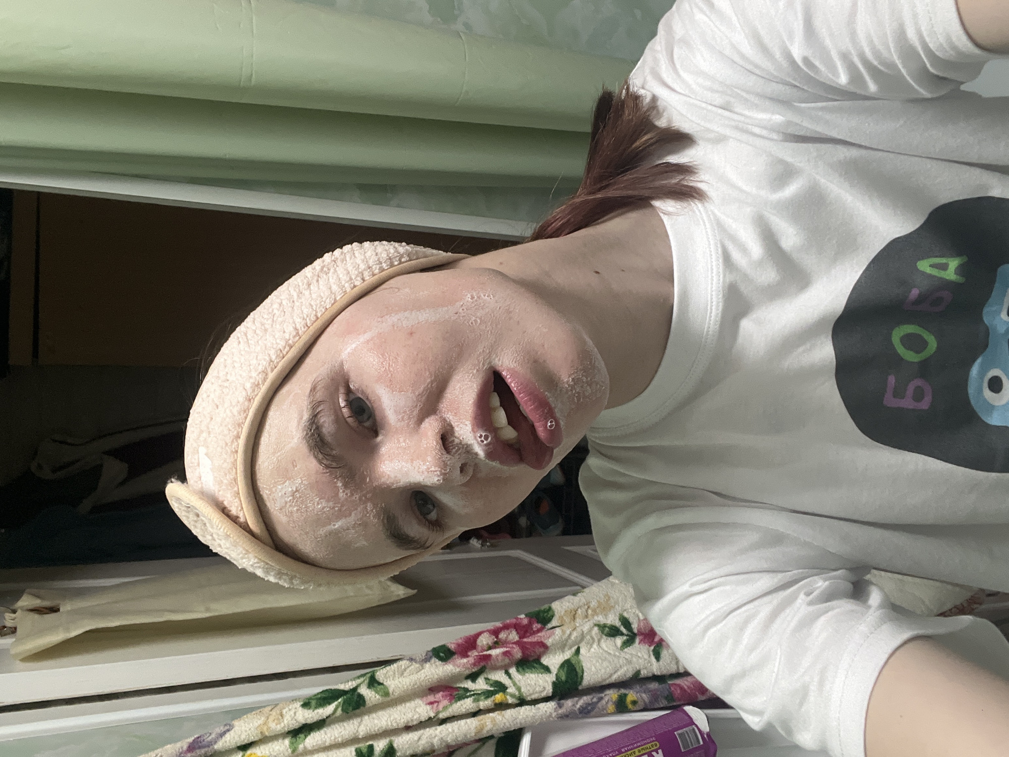 The image captures an individual, likely female, in an indoor domestic setting, reportedly in Chaikovsky, Russia. The person is positioned in a close-up, selfie-style shot, looking directly into the camera. Their face is largely covered with a white, foamy substance, indicative of a facial cleanser or mask, exhibiting small bubbles around the mouth, which is slightly open. A light pink, textured headband is worn to keep hair away from the face; the visible hair is reddish-brown and pulled back into a ponytail. The individual is wearing a white, short-sleeved t-shirt with a prominent dark circular graphic on the chest, displaying partially visible Cyrillic text that reads "БОБА". The background features a floral-patterned towel (white with pink and green designs) hanging on the left, partially obscuring a purple object. To the right, a light green shower curtain or similar fabric panel is visible, with a darker brown surface appearing behind the person's head. The scene is illuminated by ambient artificial light.