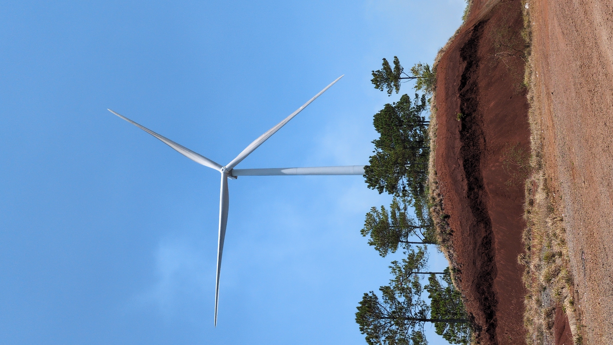 The image depicts a wind turbine against a blue sky, juxtaposed with a view of the landscape near Phát Chi, Vietnam. The right side of the image shows a dirt road and a sharply eroded embankment with layers of red soil and vegetation. The contrast between the industrial structure and the natural environment is striking. No people are visible.