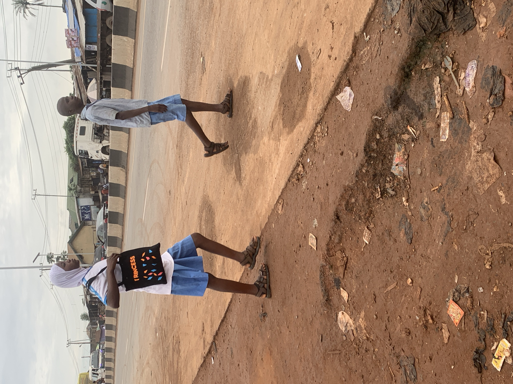 In Shagamu, Nigeria, two individuals walk along the side of a road. On the left, a young female wearing a white hijab, white shirt with blue stripes, and a blue skirt walks while carrying a black tote bag with the word "PRINCESS" printed in orange letters. She is wearing brown sandals. To her right, a young male in a light blue short-sleeved shirt and light blue shorts walks, also wearing brown sandals. They are walking on a dirt path adjacent to a road with cars and trucks in the background. The ground is a mix of dirt and debris, with scattered pieces of trash visible. The sky is overcast.