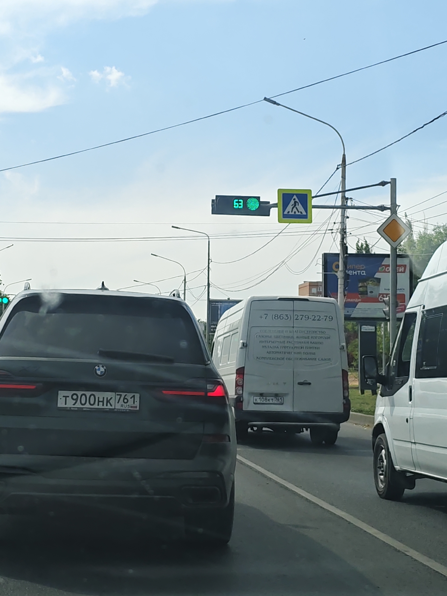 This image depicts a street scene in Novocherkassk, Russia, on a clear day with a partly cloudy sky. In the foreground, a dark-colored SUV with a license plate reading "T900HK 761 RUS" is visible from the rear. To its right, a white van with the license plate "K108KT 761" is partially visible, and further to the right, another white van is the most prominent vehicle in the frame. In the middle ground, a traffic light is illuminated green, showing the number "63" and a pedestrian crossing sign. A diamond-shaped yield sign is also visible. The environment includes overhead power lines and streetlights. The overall impression is a snapshot of traffic on a city street.