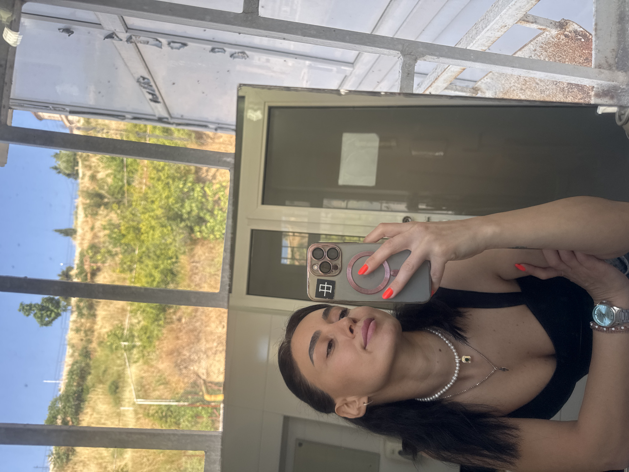 A young woman with dark hair, wearing a black top, pearl necklace, and a watch, takes a mirror selfie outdoors. The background shows a grassy hillside with trees and a building, possibly a church, on top. There are metal bars in the foreground, suggesting she is behind a window or fence. The overall scene appears to be in a city environment, likely Tbilisi, Georgia, given the architectural style and landscape.