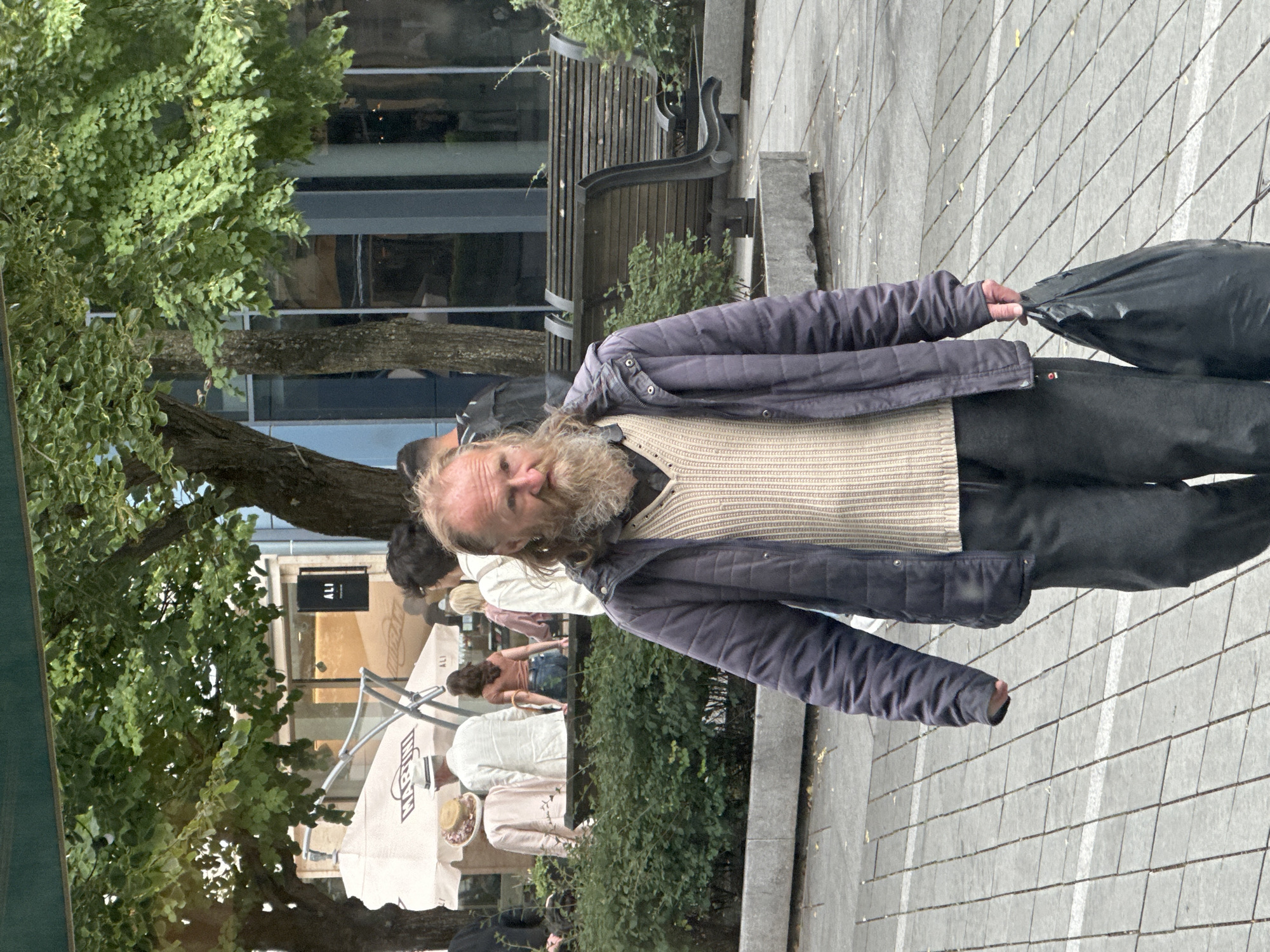 A middle-aged Caucasian man with a long, unkempt beard and disheveled hair walks towards the camera on a paved city sidewalk. He is wearing a dark blue quilted jacket over a cream-colored ribbed sweater and dark pants. In his right hand, he carries a black plastic bag. In the background, other pedestrians are visible, some standing near white umbrellas and shop fronts with visible signage, including "ALI." Behind them, a row of wooden benches sits in front of lush green bushes and trees, with the facade of a modern building visible in the distance. The scene appears to be an outdoor public space, potentially a pedestrian street or square in Kaunas, Lithuania, characterized by urban landscaping and commercial establishments.