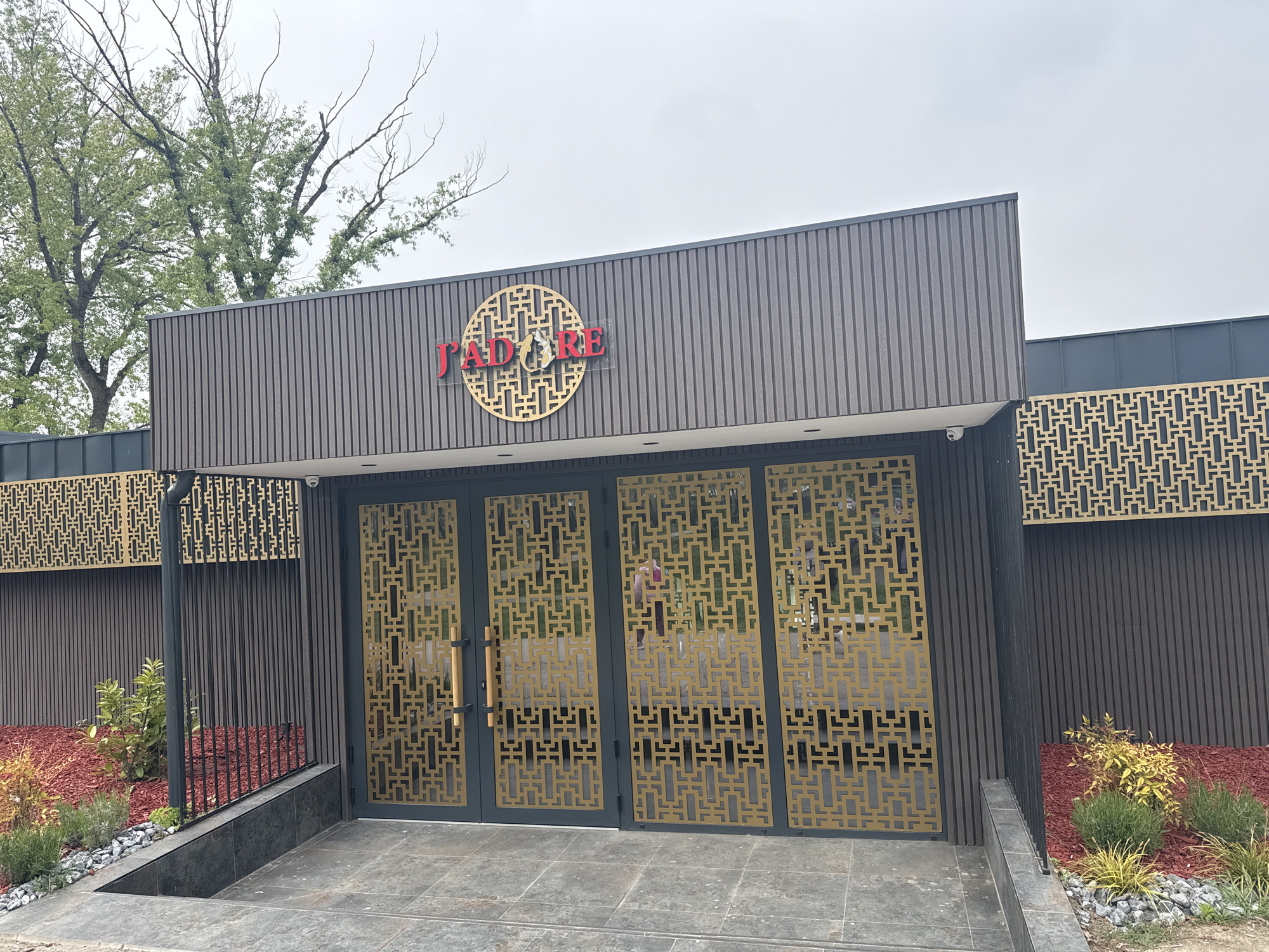 The image shows the entrance to a building with a sign that reads "J'ADORE." The building has a modern design with dark brown vertical siding and a gold geometric pattern on the doors and surrounding panels. The landscaping includes red mulch, small shrubs, and decorative stones. The sky is overcast, and there are trees visible in the background. The building's location is in the city of Unknown, Unknown.