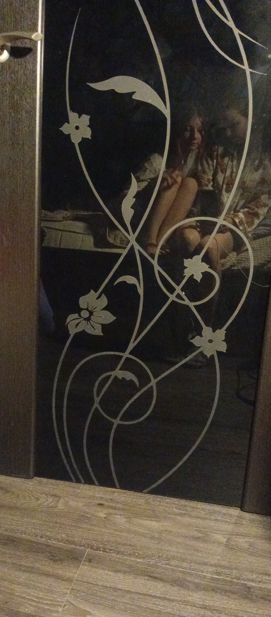 The image depicts a dark-colored panel with an ornate, light-colored floral design. The panel is framed by a brown material. The background is dark. Reflected in the panel is a person with their legs visible and wearing a patterned robe. The panel is set against a wood-look floor. The location appears to be indoors, with the scene being a reflection.