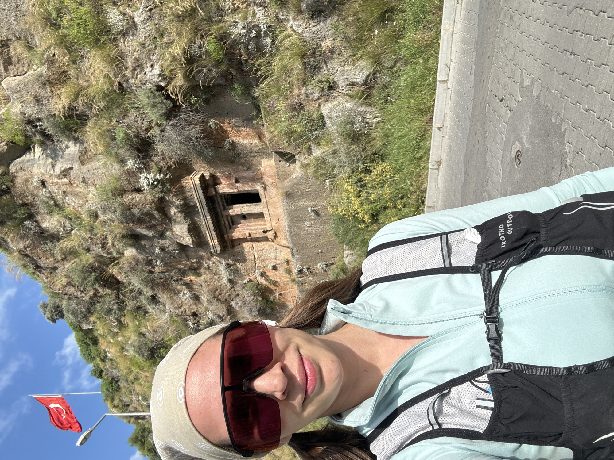A medium shot features a woman wearing a light-colored headband, sunglasses, and a light-colored jacket with a black and white backpack. She is facing the camera with a slight smile.
Behind her, a hillside is visible, with a square-shaped tomb carved into the rock face. Vegetation grows on the hillside and the surrounding area.
To the left, a Turkish flag is visible against a blue sky with scattered clouds.
The scene appears to be outdoors, likely in a tourist or historical site, as the tomb suggests. The location is the city of Fethiye, Turkey. A paved walkway is on the right.