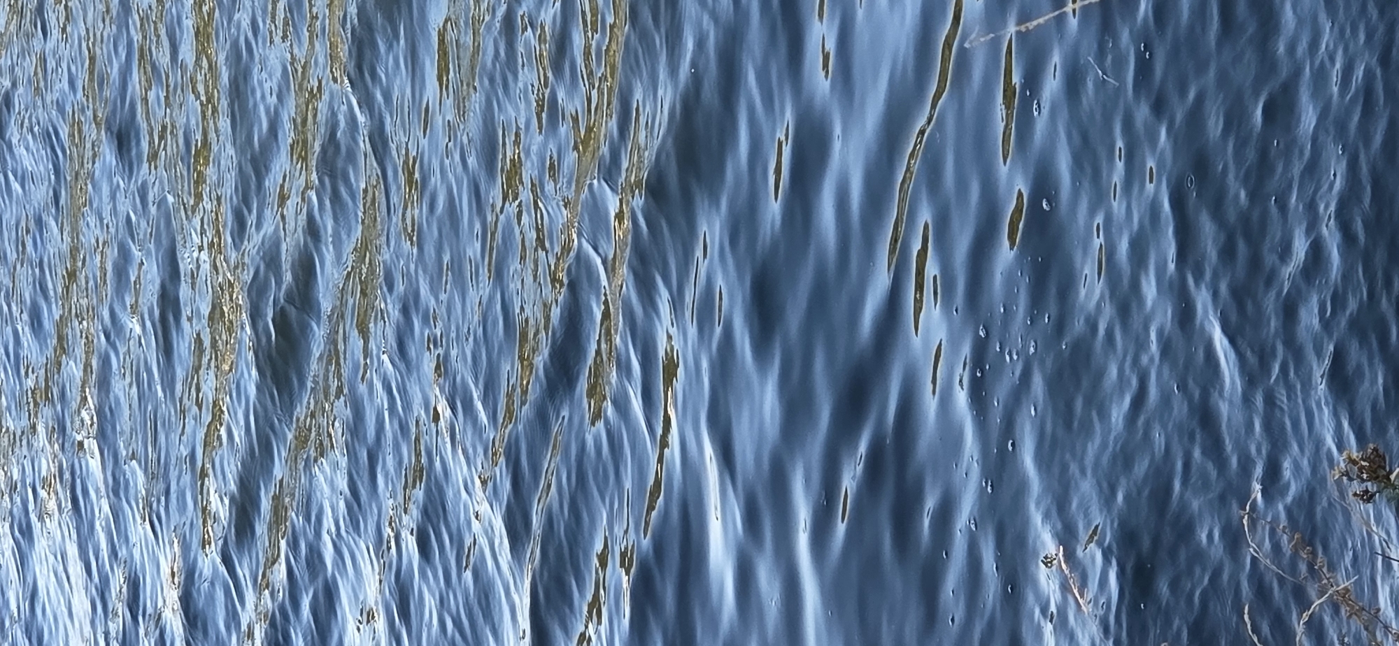 This is a close-up, vertical, eye-level shot of the surface of a body of water, possibly a river or canal, in Strasbourg, France. The water is dark blue with subtle ripples creating varying shades of blue and highlights from sunlight reflecting off the surface. There are small, scattered air bubbles visible, particularly in the lower left quadrant of the image. In the very bottom left and right corners, there are glimpses of dry, brown vegetation, suggesting the edge of the water. The scene is devoid of any people or man-made structures, focusing solely on the natural texture and movement of the water.
