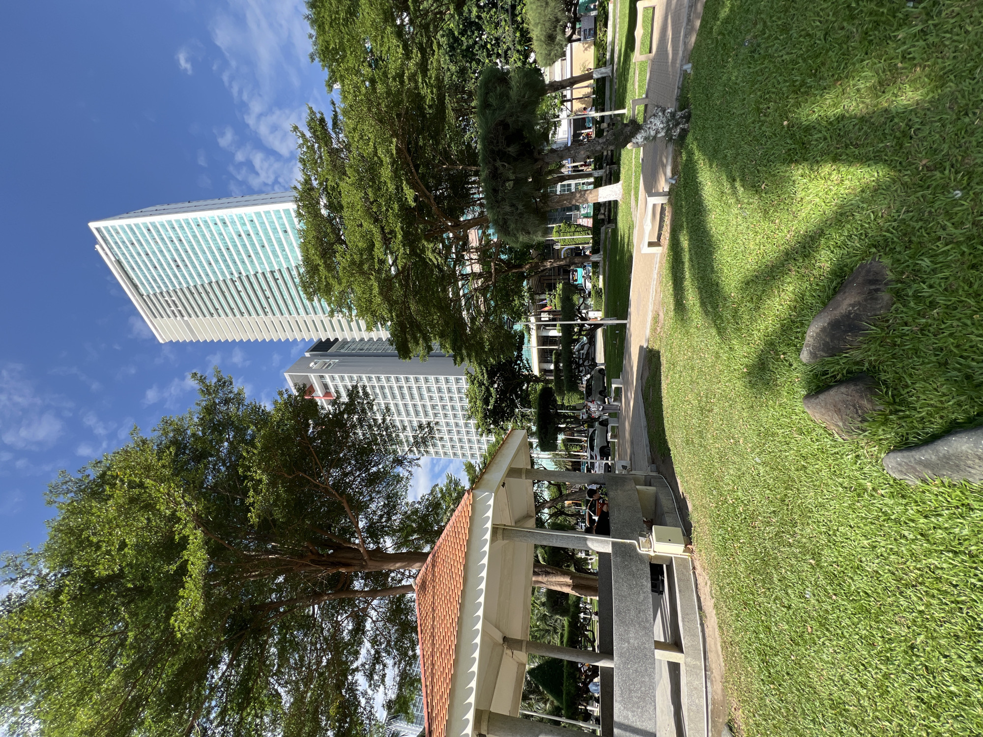 The image depicts a public park in Nha Trang, Vietnam, on a bright, sunny day with a clear blue sky and scattered clouds. In the foreground, a well-maintained lawn is visible, with two dark, rounded stones placed on it. Behind the lawn, there is a covered pavilion with a red tiled roof supported by pillars. This pavilion appears to be made of concrete and stone, and there are people seated inside and around it.
Beyond the pavilion, a paved walkway runs through the park, with several white benches lining its edge. Lush green trees with dense foliage are scattered throughout the park, casting shadows on the grass. In the background, two modern, tall buildings rise into the sky. The building on the right is particularly striking, with its white facade and rows of blue-tinted windows, appearing to be a hotel or apartment complex. The building to its left is also a multi-story structure, with visible balconies and windows. Several cars are parked along a street or driveway visible between the pavilion and the buildings. The overall scene suggests a pleasant urban recreational space.