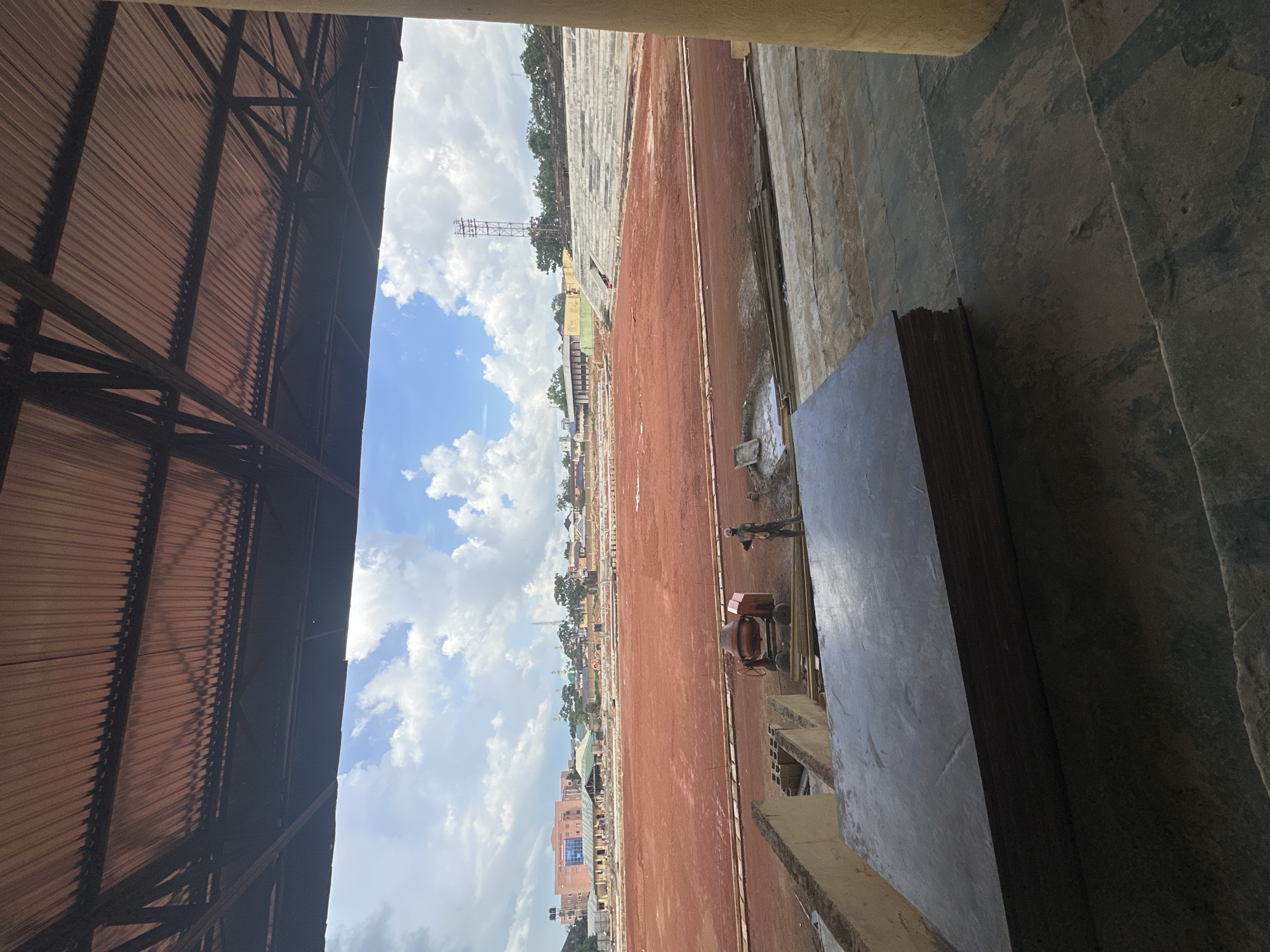 The image depicts the ongoing construction of a sports stadium in Jos, Nigeria. The stadium features a red dirt field, surrounded by concrete seating structures. Construction equipment, including a cement mixer, and building materials like cinder blocks and plywood sheets are visible on site. A lone construction worker stands near the cement mixer. In the background, urban buildings and trees dot the landscape under a cloudy sky.