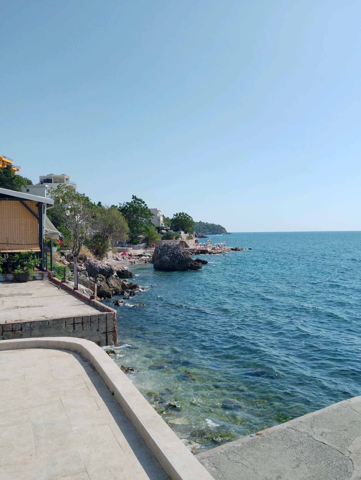 This image depicts a sun-drenched scene of the coastline in Sutomore, Montenegro. In the foreground, a textured concrete walkway with a curved edge leads the eye towards the sea. To the left, a wooden structure with a thatched roof and bamboo blinds is partially visible, adorned with greenery and red flowers. The rocky shoreline is dotted with people sunbathing and relaxing under umbrellas on a small beach area. Buildings, some with white facades and balconies, are situated on the hillside behind the beach, nestled amongst lush green trees. The expansive, clear blue sea stretches out to the horizon under a bright, cloudless sky. Waves gently lap against the rocks, and the water is transparent enough to reveal submerged stones and vegetation. The overall impression is one of a peaceful and inviting coastal resort.