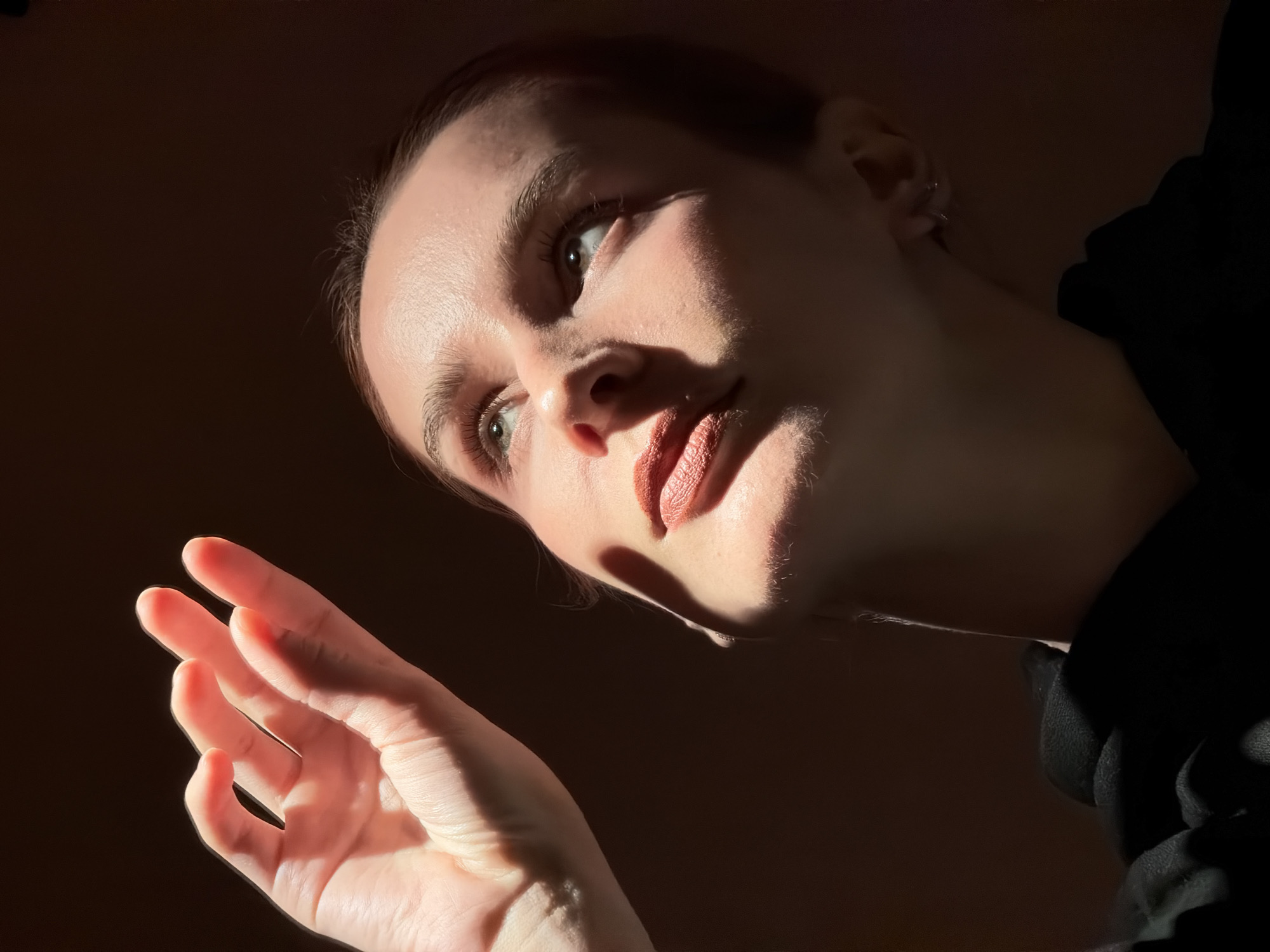 A close-up, low-angle portrait features a fair-skinned woman, likely indoors, with a dramatic lighting effect. She is looking slightly upwards and to the left, her expression neutral. Her makeup is minimal, with noticeable lipstick. Her hair appears to be pulled back.
The woman's hand is visible in the lower-left corner, palm facing upwards, fingers slightly spread. The background is a dark, indistinct color.
Based on the prompt, this image could have been taken in the city of Chaikovsky, Russia. However, there are no identifiable features in the image to confirm this location.