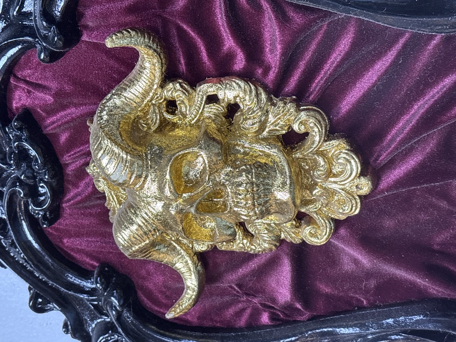 A golden, ornate skull relief with prominent horns is centrally displayed against a deep purple velvet background, partially framed by a glossy black, heavily sculpted border. The skull features textured golden surface detail, including eye sockets, nasal cavity, and teeth, with elaborate baroque-style scrollwork integrated into its design, particularly around the jawline and extending towards the base of the horns. The horns curve upward and outward, mimicking animal horns. The velvet fabric shows subtle folds and a soft sheen. This decorative arrangement is located indoors in Nam Hoa Lư Ward, Vietnam. No people, actions, or overt interactions are present in the scene.