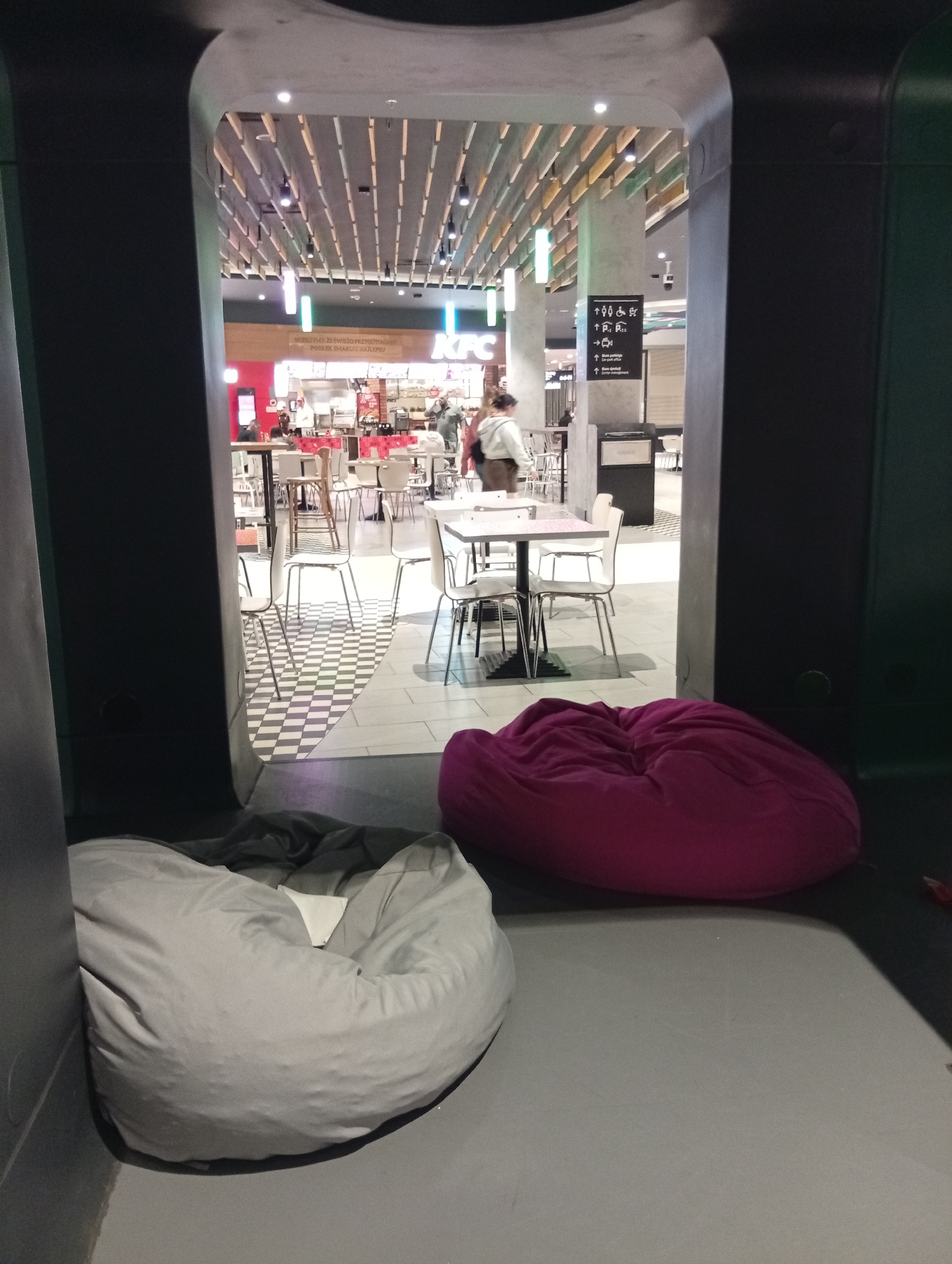 In Wrocław, Poland, at a KFC restaurant, two large beanbag chairs are visible in the foreground. A gray beanbag is on the left, and a magenta beanbag is on the right. Beyond the beanbags, a KFC restaurant interior is visible with tables and chairs. Several people are seated at tables, and staff members are visible behind the counter. The floor has a black and white checkered pattern. A sign with pictograms indicating directions to facilities like restrooms and elevators is mounted on a pillar. The restaurant has a modern ceiling with decorative wooden slats and lighting fixtures.