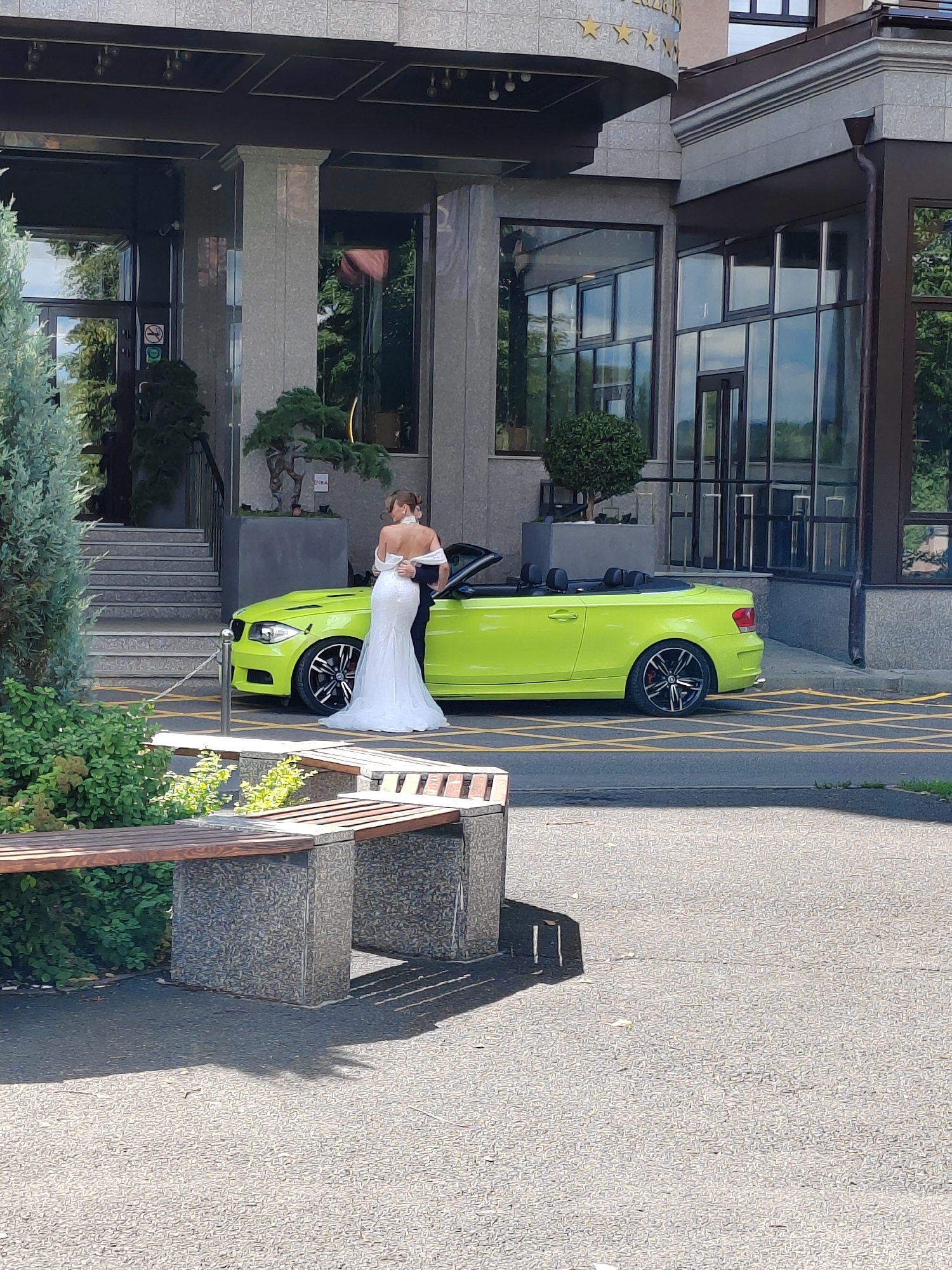 A couple, likely a bride and groom, are embracing in front of a bright yellow convertible car parked on a street. The woman is wearing a white wedding dress with an off-the-shoulder design, and the man is in a dark suit. The car is parked in front of a modern building with large glass windows and stone accents. In the foreground, there is a wooden bench with stone supports, and the ground is paved with asphalt. The scene appears to be a sunny day, with shadows cast on the ground from the bench and the building. This appears to be the building located at the intersection of Krasnaya Gorka Street and Severnaya Street in Kemerovo, Russia.