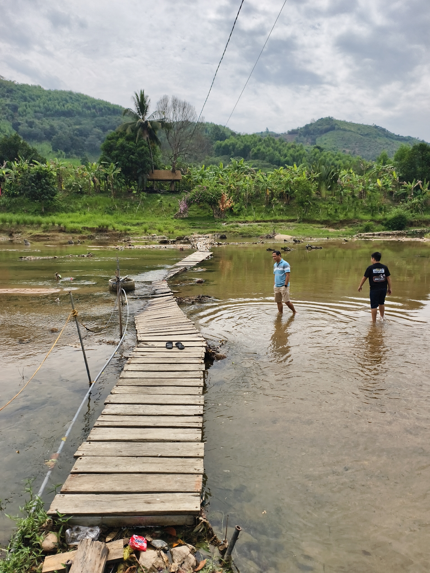The photograph captures a rural scene in Nam Khanh Vinh, Vietnam. A makeshift wooden bridge crosses a shallow, murky river. The bridge is constructed from planks and is partially submerged in the water. Two men are wading through the river next to the bridge. One man is wearing a blue striped shirt and khaki shorts, while the other is in a black t-shirt and dark shorts. On the bridge are a pair of sandals. Litter and debris are visible at the edge of the bridge. The background features lush green vegetation, including trees and banana plants. A small pavilion-like structure is situated among the trees. Hills and a cloudy sky form the backdrop. Overhead, wires stretch across the scene, possibly electrical or utility lines.