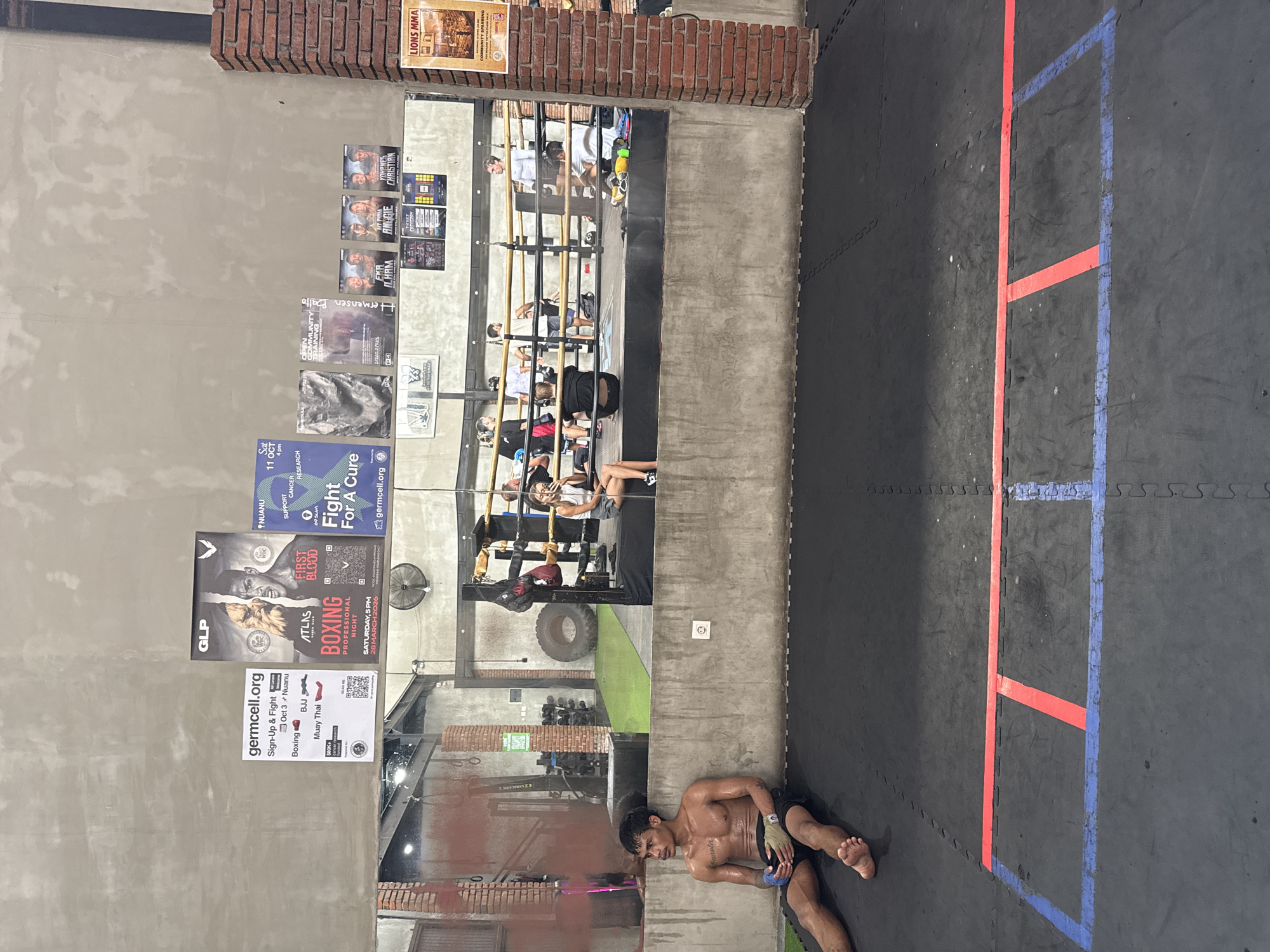 This media file captures an indoor combat sports training facility, likely in Mengwi, Indonesia, with a raw, industrial aesthetic featuring exposed concrete walls and some brickwork. The floor is covered with black interlocking mats, marked with red and blue lines that delineate a training area, possibly for a boxing or MMA ring.
In the foreground, a shirtless, sweaty man with dark hair is seated on the mat, resting. He appears to be taking a break from physical exertion, holding a small object in his hand, possibly a water bottle or a phone. His expression is neutral, suggesting fatigue.
Dominating the left wall is a large mirror reflecting several individuals, who appear to be engaged in a training session or observing one. Some are standing, others seated or positioned on the floor, suggesting active participation in a class or sparring. Behind the reflection, gym equipment like a large tire and dumbbells are visible.
The concrete wall space is extensively used for advertising and announcements. Numerous posters are affixed, promoting various combat sports events. Visible text includes "LIONS MMA COMMUNITY SPAR," and posters detailing "ATLAS BOXING FIRST BLOOD PROFESSIONAL NIGHT" scheduled for "SATURDAY, 5PM 28 MARCH 2026." Another poster advertises "germcell.org Sign-Up & Fight" for "Boxing, BJJ, Muay Thai" on "Oct 3 Nuanu." A "Fight For A Cure" event supporting cancer research, also associated with "germcell.org," is announced for "Sat 11 OCT 4 pm" in "NUANU." Other posters feature names like "Ballo termensen," "EKA ILHAM," "ANGGIE," and "YOHANES CHRISTIAN," likely promoting fighters or participants.
The lighting is artificial, suggesting the image could have been taken at any time of day inside the gym. The overall atmosphere points to a dedicated and active training environment.