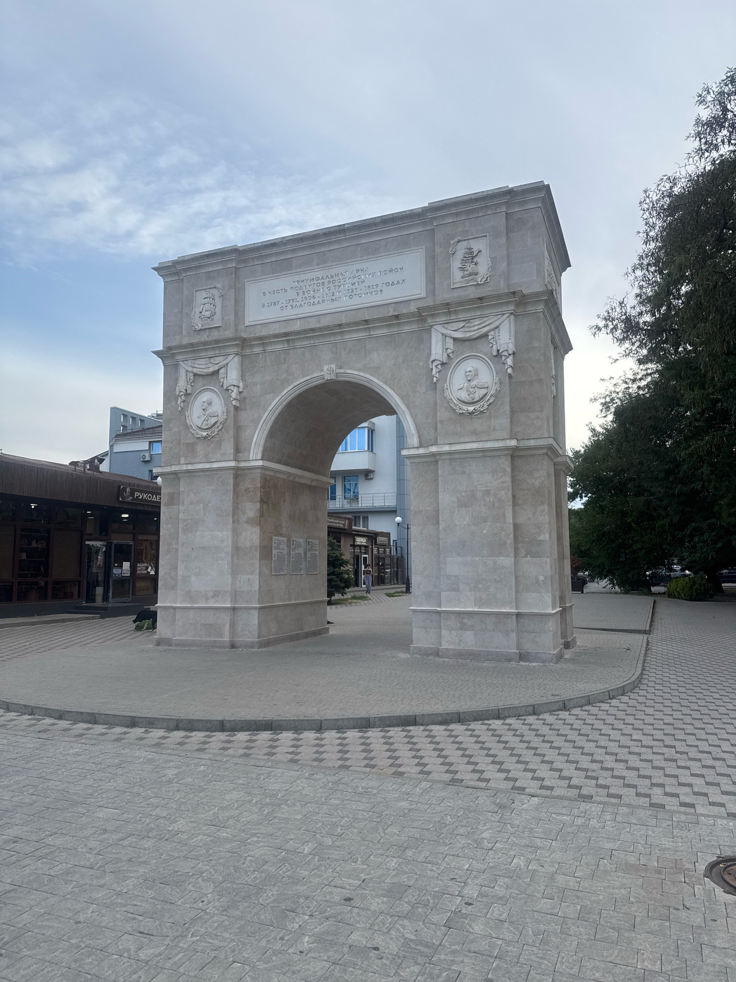 The image shows the Triumphal Arch in Anapa, Russia, a monument dedicated to the Russian soldiers who fought in the Russo-Turkish War. The arch is constructed of light-colored stone and features bas-reliefs and inscriptions. In front of the arch is a paved square with geometric patterns. To the left of the arch, there are buildings with shops, and to the right, a large tree. The sky is partly cloudy. There are no people clearly visible in the foreground.