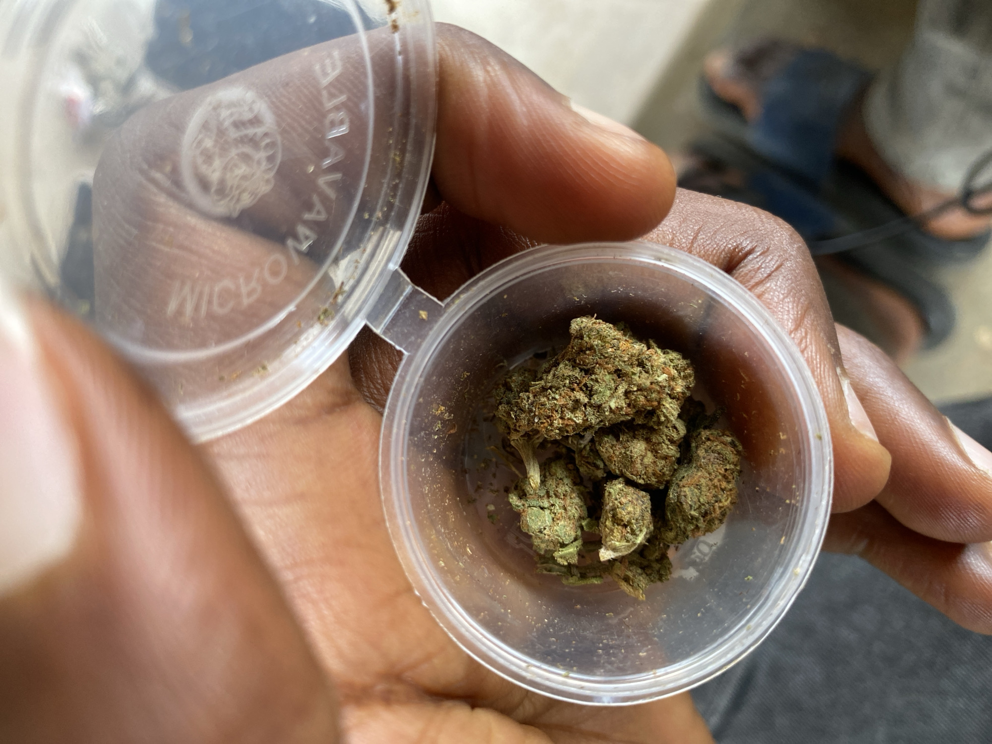 A close-up, top-down shot features a dark-skinned adult hand holding a small, transparent plastic container filled with several green and orange-brown marijuana buds. The container's lid is open and partially attached on the left, displaying embossed text that appears inverted and reflected. Visible text on the lid includes "MICROWAVABLE" and another phrase, possibly "DISHWASHER SAFE," along with a circular, intricate logo or pattern.
The focus is on the hand and the contents of the container. The background is blurred, showing soft shapes and colors. In the upper right portion of the blurred background, a person's lower legs and feet wearing dark-colored sandals are visible, suggesting a casual, relaxed setting, possibly indoors or on a patio. The lighting appears bright, indicative of daytime. The image captures the moment of holding or inspecting the cannabis, with the location context provided as Jos, Nigeria.