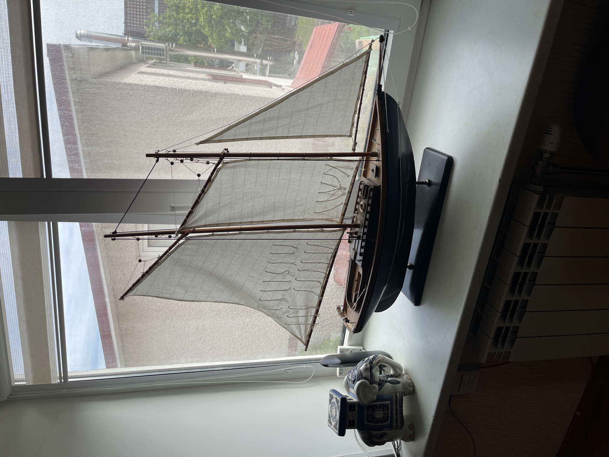 The image depicts a model sailboat on a windowsill. The sailboat is rigged with two white sails and dark wooden masts. It is mounted on a black display stand. Beside the boat is a blue and white ceramic elephant figurine. The window behind the boat is partially obscured by a white roller blind, with the view showing rooftops and trees, suggesting a residential area. A radiator is visible along the windowsill. The image is taken from a high angle, looking down on the scene.