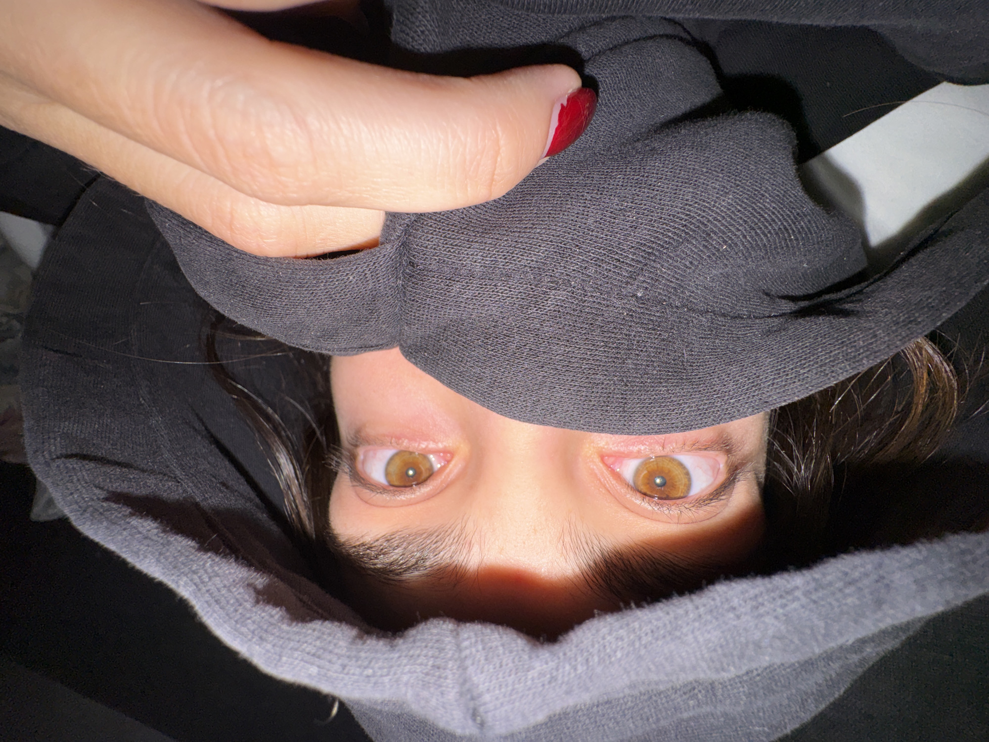 A person with brown eyes is seen in a close-up, upside-down view, peering out from beneath a grey hooded sweatshirt. A hand with red nail polish is visible at the top left of the frame, holding the hood. The background is dark and obscured. There is no visible information to confirm the location is Mexico City, Mexico.