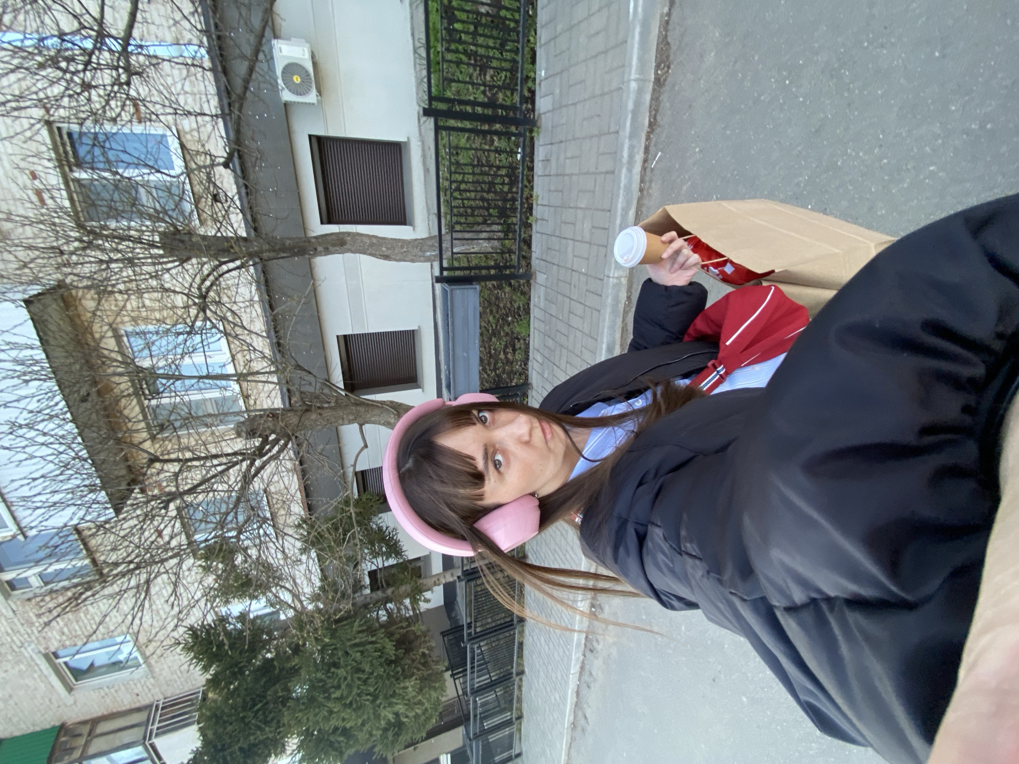 A young woman wearing pink headphones and a black jacket stands outside, holding a coffee cup and a brown paper bag. Behind her is a light-colored building with windows, dark shutters, and an air conditioning unit. A leafless tree is visible in front of the building. The scene appears to be in an urban area, likely a street in Votkinsk, Russia, as indicated by the location. The woman is looking at the camera.