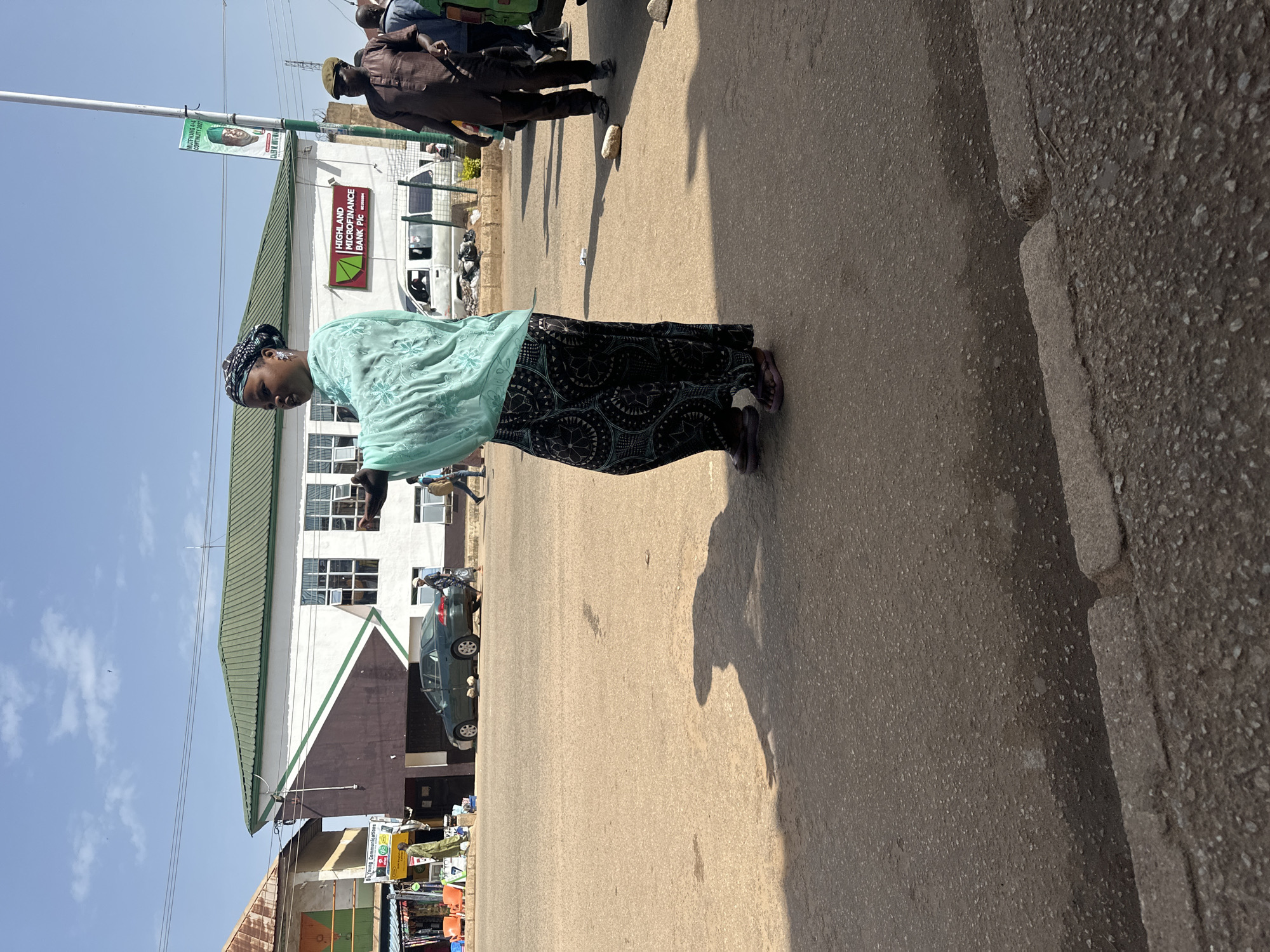 In the city of Bukuru, Nigeria, a woman wearing a light blue garment and a dark patterned skirt stands on a paved road. She is looking to her left, with her right arm extended. Behind her, a white building with a green roof and a dark brown diagonal stripe is visible. The building has multiple windows and a sign that reads "HIGHLAND MICROFINANCE BANK Plc". A green sedan is parked in front of the building. Other people are present in the background, including a man in a brown outfit and a cap walking to the right. The road surface is asphalt, with some debris and stones scattered on it. The sky is clear and blue, with a few wispy clouds. The lighting suggests it is daytime.