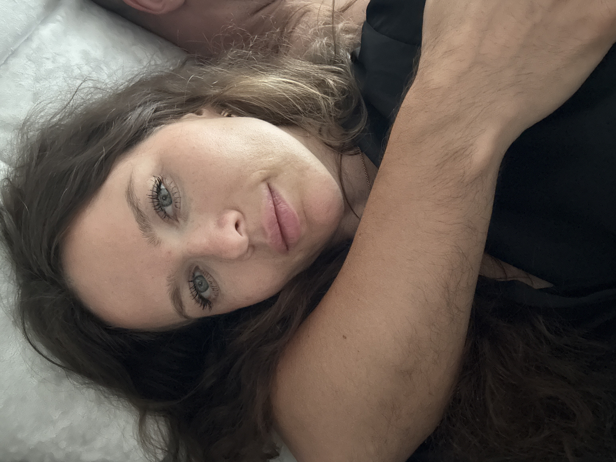 A woman with long, dark brown hair is shown in a close-up selfie, lying down. She has blue eyes, light skin with freckles, and her lips are slightly parted. She is wearing a black top. Her left arm is wrapped around her body, partially obscuring her chest, and her right arm is visible across the foreground. The background appears to be a textured white surface, possibly a pillow or blanket. The lighting is soft and natural. No specific location in a city can be determined from the image.