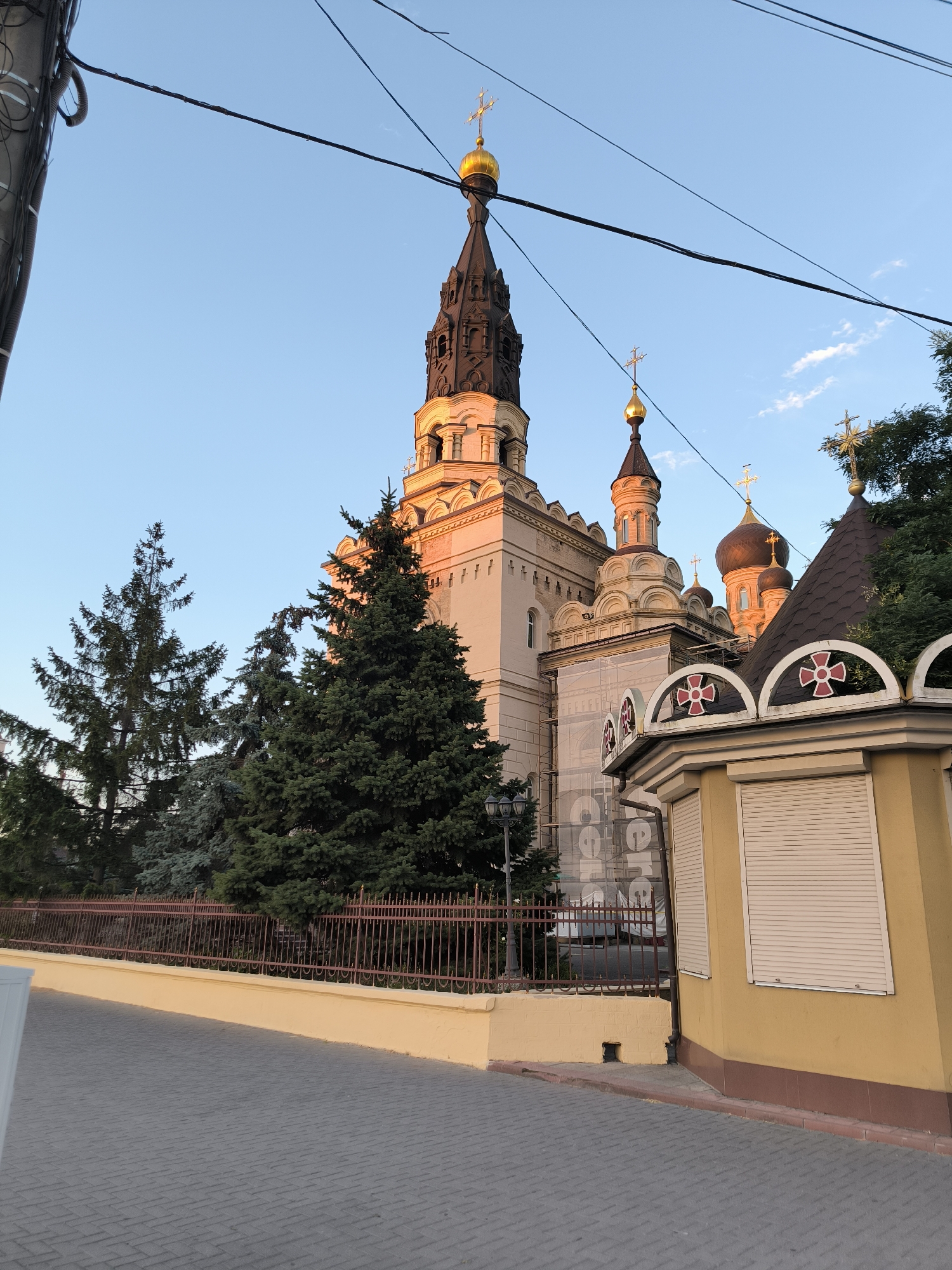 The image captures a portion of a large, ornate building, likely a church, bathed in the warm light of the setting sun. The architecture features multiple domes and spires topped with golden crosses, characteristic of Eastern Orthodox churches. The main structure is a light-colored stone building with decorative elements and arched windows. In the foreground, a paved walkway leads towards the building, bordered by a low yellow wall and a dark metal fence. Several evergreen trees, including tall pine trees, are visible behind the fence, adding a touch of nature to the urban landscape. A small, yellow, shed-like structure with a roll-down shutter stands to the right, partially obscuring the main building. Overhead, power lines are strung across the clear blue sky, with a few wisps of clouds. The scene appears tranquil and serene, with no people visible.
This building is likely the St. Nicholas Cathedral in Mykolaiv, Ukraine. The cathedral, dedicated to Saint Nicholas, is a prominent landmark in the city and is known for its impressive architecture and golden domes. The surrounding environment, with the paved walkway, fence, and trees, is consistent with the area around the cathedral. The presence of the power lines suggests an urban setting within Mykolaiv. The warm lighting indicates the photograph was taken during either sunrise or sunset.