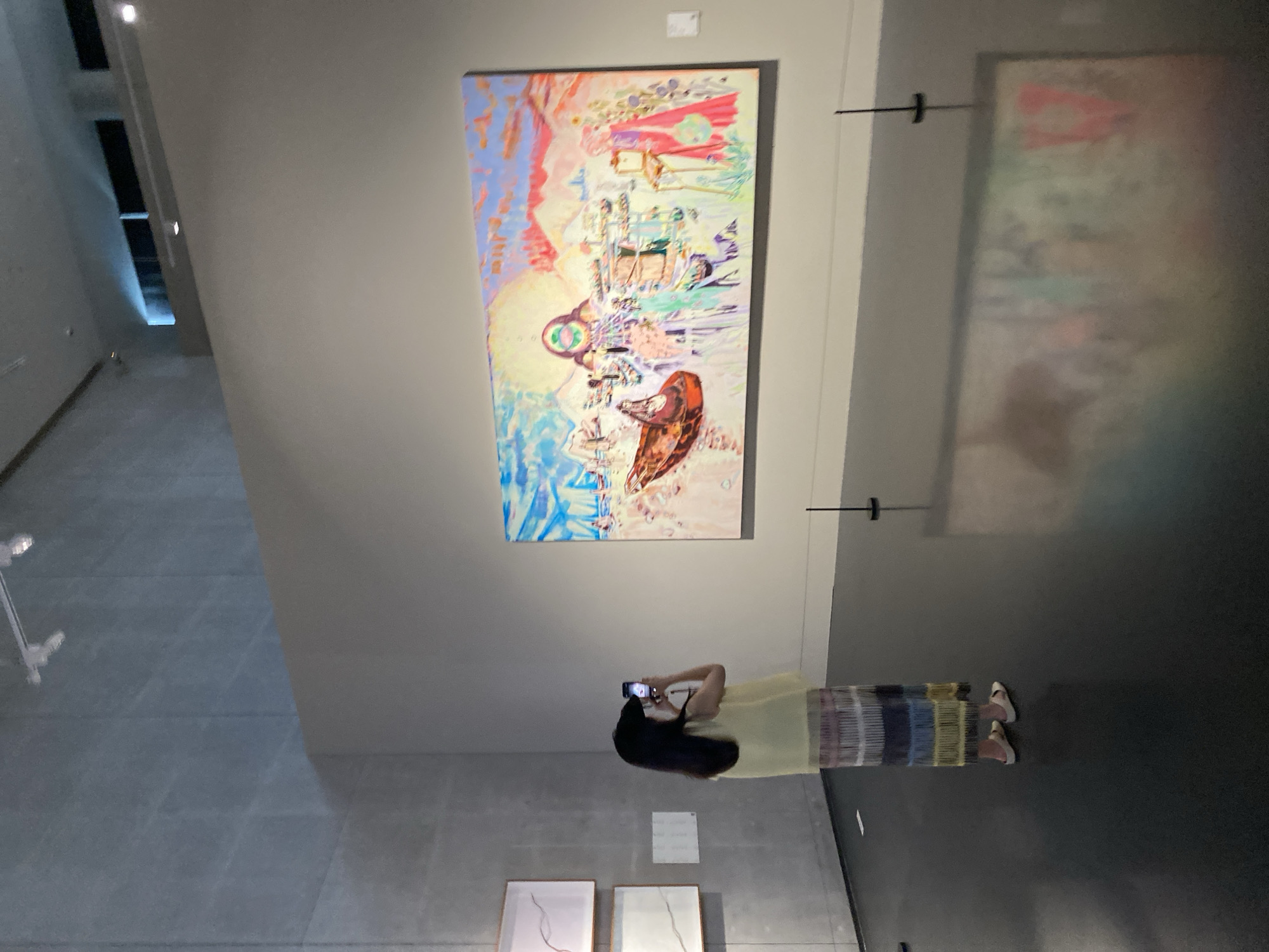 A person, viewed from the rear, stands in an art museum in Jinniu District, China, photographing a large, colorful abstract painting on a gray wall with a smartphone. The painting depicts a surreal landscape with elements that suggest figures, structures, and a boat. The person wears a pale yellow sleeveless top and a long skirt with horizontal stripes of various colors, including purple, blue, and yellow, and white sandals. In the background to the left, two framed pieces of art with white backgrounds and thin dark lines are mounted on a concrete wall. The museum floor is dark and reflective. The lighting appears to be from overhead fixtures.