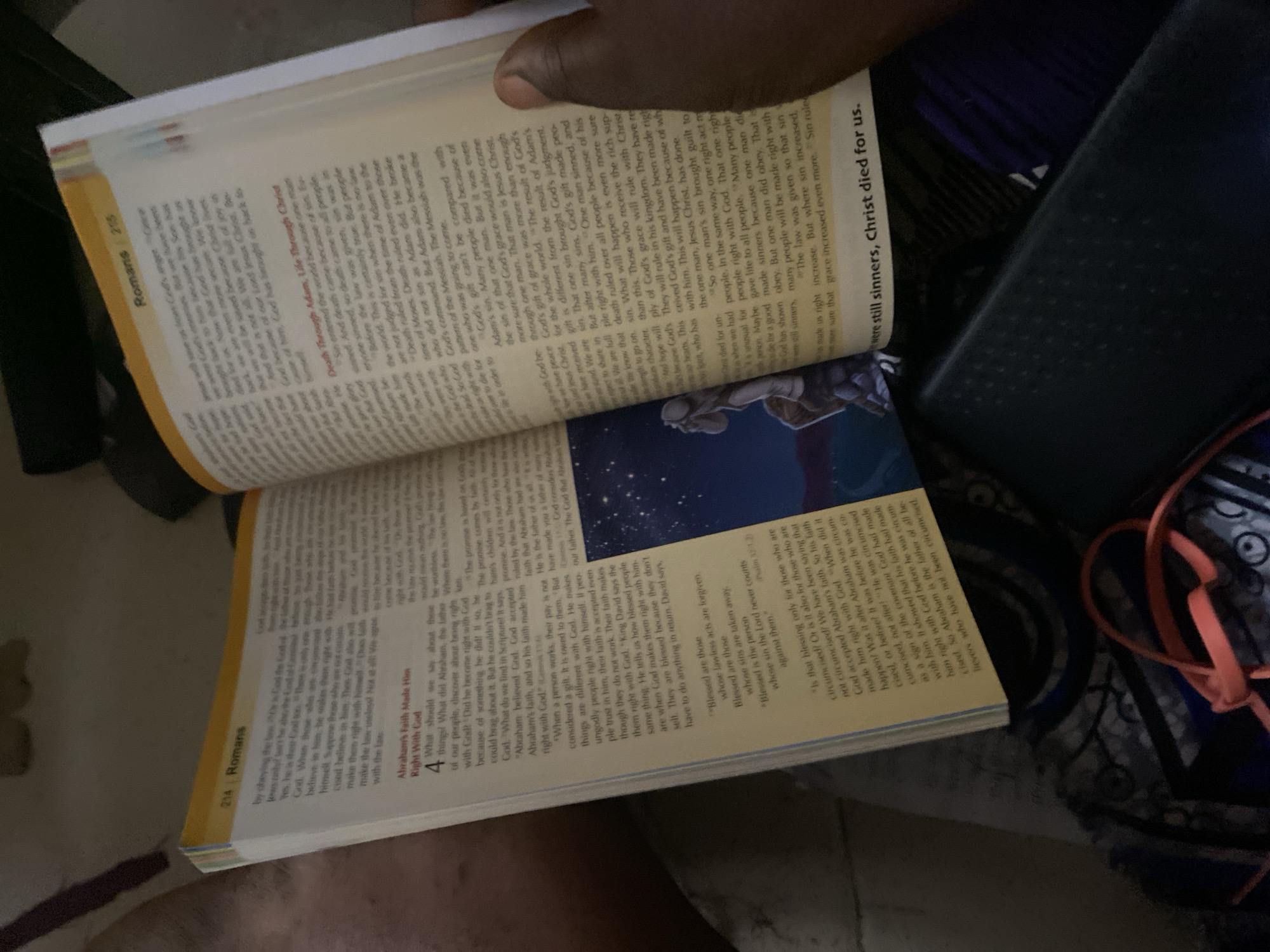 The image shows an open Bible, with text from the book of Romans. The left page contains Romans 4:1-5, discussing Abraham's faith. The right page contains Romans 5:6-8, discussing Christ's death for sinners. The Bible is placed on a surface that appears to be a dark, textured object, possibly a laptop or a textured table. The lighting is dim, with some shadows cast across the page. There is a hand visible on the right side of the image, holding the edge of the right page. The overall scene is a close-up of the Bible, with the focus on the text. There is no indication of the location in the city of Jauro Yinu, Nigeria.