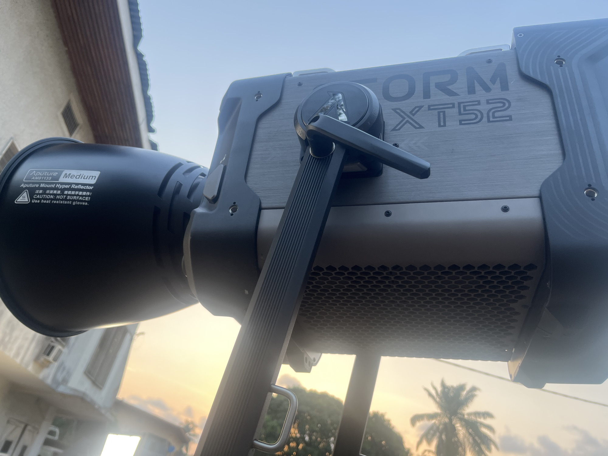 The image shows an Aputure LS 600d Pro light with a Hyper Reflector attached. The side of the light's body reads "Storm XT52." The reflector has a warning label about the surface getting hot and advises the use of heat-resistant gloves. The light appears to be set up outdoors, based on the background view of a building, palm tree, and a sky indicating either dawn or dusk. Based on the appearance of the sky and the presence of palm trees, the location might be Lagos, Nigeria.