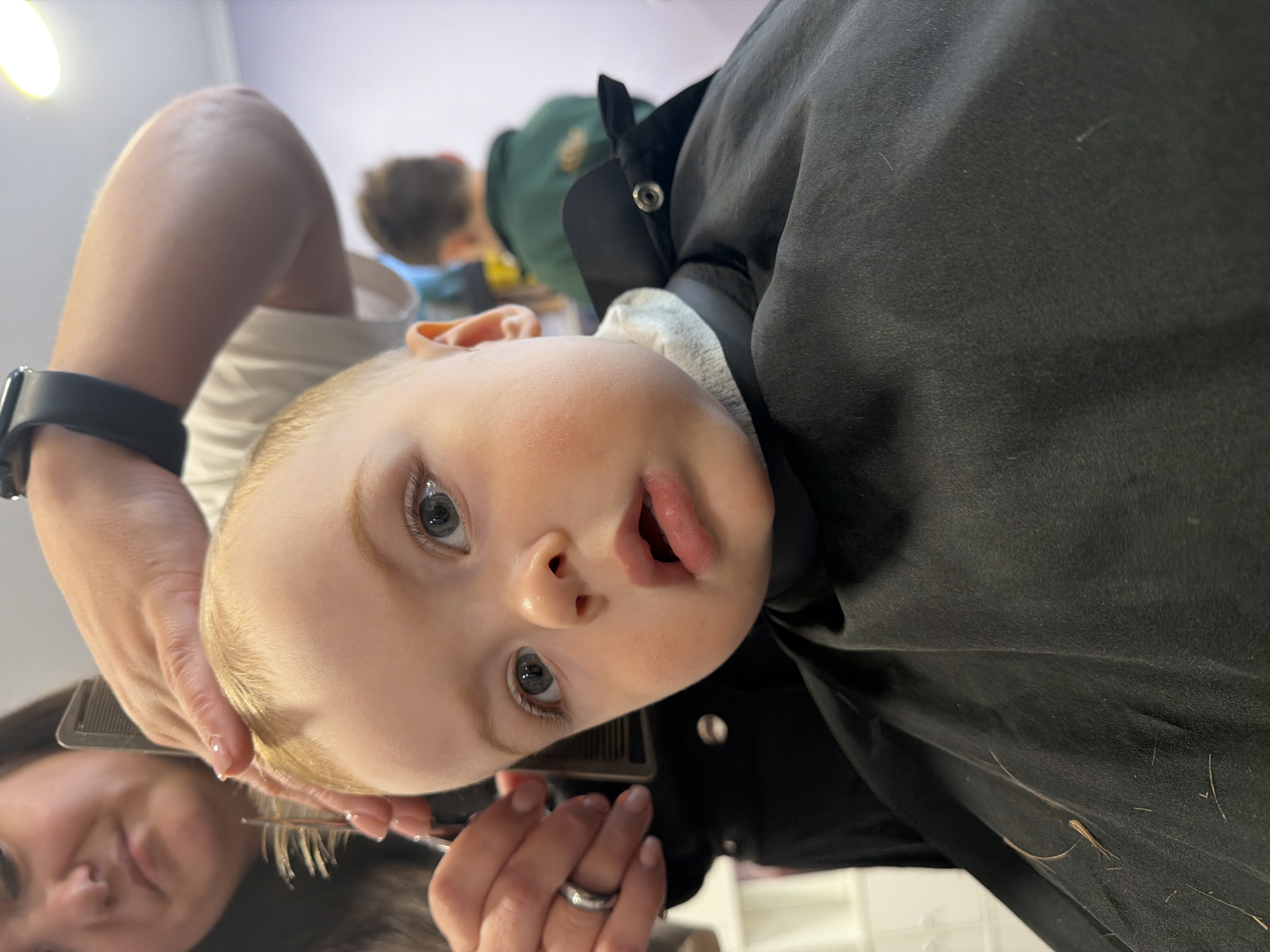 A fair-skinned, blonde-haired toddler, with blue eyes and an open mouth, is getting a haircut. The child is wearing a black haircut cape. A person, visible from the shoulders up, is using scissors and a comb to cut the child's hair. The person is wearing a black smartwatch on their left wrist and a ring on their left ring finger. Another person is visible in the background. The setting is indoors, likely a home or a barbershop, with a white shelf visible in the background. There is no specific indication or information that would indicate this event to be in Votkinsk, Russia.