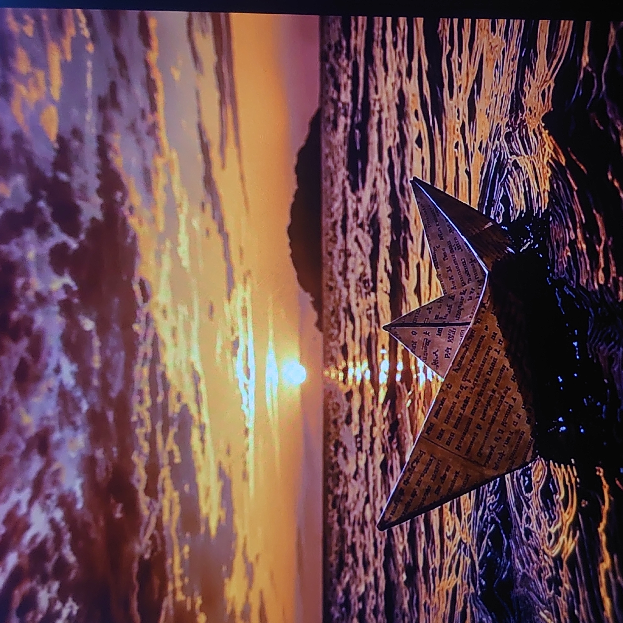 The image depicts a paper boat floating on the water during sunset. The sky is filled with dramatic clouds in shades of purple and orange, with the sun setting on the horizon, casting a golden reflection on the water. The water itself appears choppy, with waves creating ripples and highlighting the sunset's colors. The paper boat is made from newspaper and is positioned in the foreground, partially submerged in the water. In the background, a silhouetted island can be seen against the vibrant sky. There are no people or animals present in the scene. This image was captured in the city of Belorechensk, Russia.