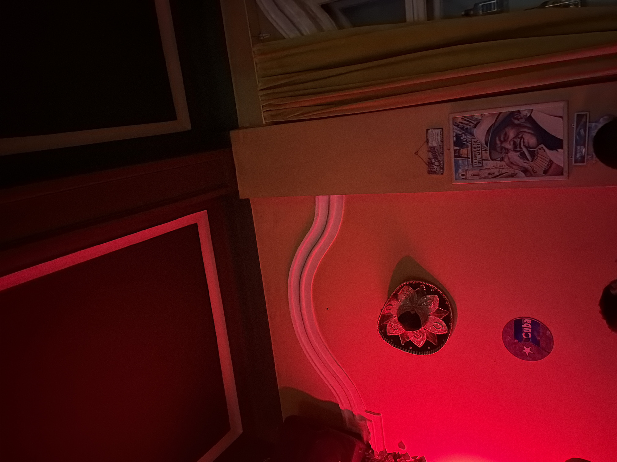 The image shows a dimly lit interior, possibly a bar or restaurant in Sofia, Bulgaria, given the Cuban-themed decorations. The room has a strong red hue. On the wall, there's a framed poster or print of a smiling man with a cigar, likely of Cuban origin. Nearby, a souvenir plate or sticker with "Cuba" written on it and a star is visible. A sombrero-shaped decoration is also on the wall. The room's architectural details include a decorative curved molding and a framed window or artwork with a black background and light trim. A curtain or awning is present near the top of the frame. The overall atmosphere suggests a themed establishment.