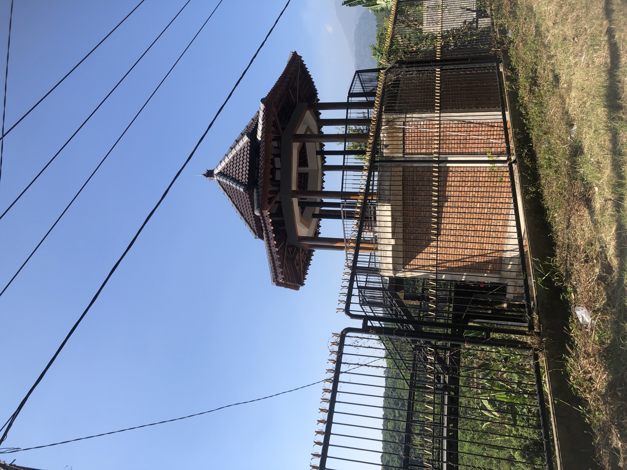 The image depicts an outdoor scene in Sukosari, Indonesia, featuring a hexagonal gazebo with a brown tiled roof and wooden pillars. The gazebo is surrounded by a black metal fence with decorative elements. To the left of the gazebo, a metal staircase leads up to a platform. The gazebo and its surroundings are set against a clear blue sky. In the background, lush green hills and mountains are visible, suggesting a rural or semi-rural setting. Power lines are strung across the upper portion of the image, and the ground in the foreground is covered with dry grass and some litter. The overall scene is one of a tranquil outdoor structure in a natural environment.