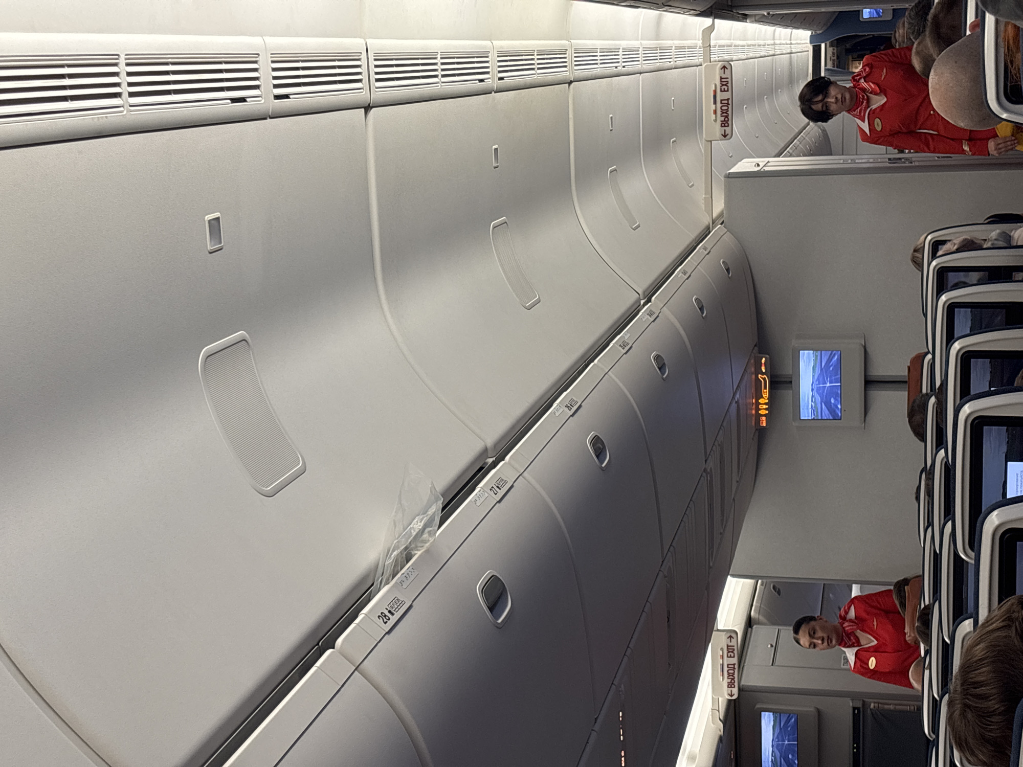 The image depicts the interior of an airplane cabin, oriented vertically. Several passengers are seated in rows of blue-upholstered seats, with the backs of their heads visible. A flight attendant in a red uniform with a red scarf is standing in the aisle, looking towards the camera. Above the passengers' heads, the characteristic curved ceiling of an aircraft is visible, featuring overhead compartments and air vents. On the wall of the cabin, an illuminated sign with Cyrillic text and an arrow points to the right, reading "ВЫХОД EXIT". Small television screens are embedded in the seatbacks. The overall scene suggests the inside of a commercial airliner, likely during a flight or boarding process, given the presence of the flight attendant and seated passengers. There are no obvious indications of the time of day or weather outside. The location context provided is Khimki, Russia, which might imply the flight is departing from or arriving at an airport in that region.