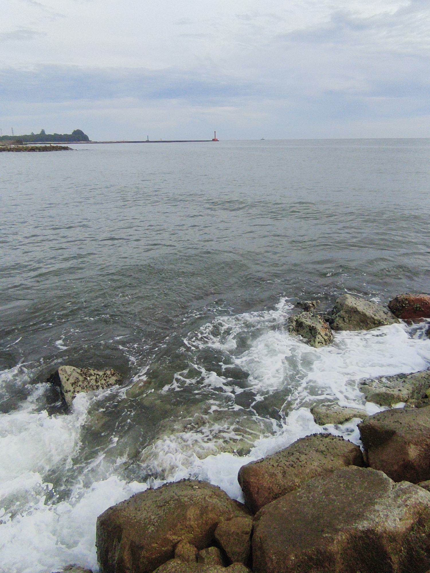 The image depicts a rocky coastline with the ocean in the foreground. Waves crash against the rocks, creating white foam. In the distance, a long pier extends into the sea, with a red lighthouse visible at the end of it. The sky is overcast, with a few clouds scattered across it. The general setting appears to be a harbor or a breakwater area.
This scene is likely located at the Ujong Blang beach area in Lhokseumawe, Indonesia. Lhokseumawe is a port city in Aceh province, known for its industrial activities, including oil and gas. The presence of a pier and lighthouse suggests a connection to maritime activities or the protection of the coastline from the sea. The rocky nature of the shore is typical of many coastal areas in the region.