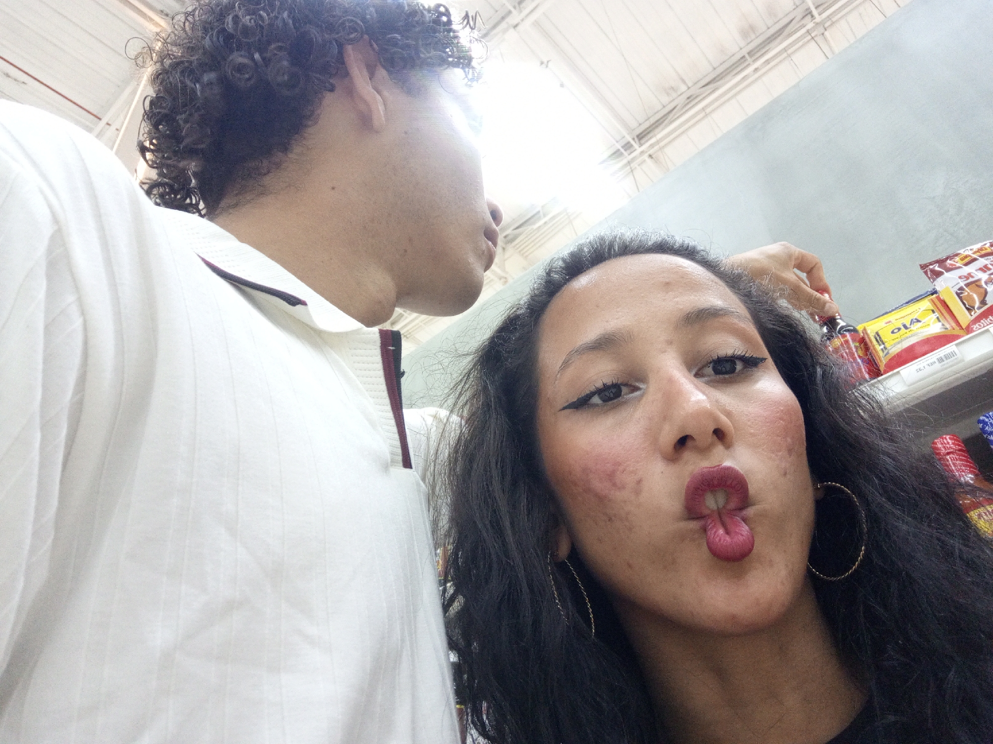 Two people are in the photo. A person with dark curly hair is on the left wearing a white collared shirt with thin red and black stripes along the collar and shoulder. The person is angled to the left of the photo, showing their side profile and neck. To the right of this person is a person with long dark hair, gold hoop earrings, and winged eyeliner. Their face is oriented towards the camera and their lips are pursed into a pout. They appear to be in a store, based on the shelves of products visible behind the woman's head. The shelf displays bottles, sauces and snacks. The top of the image reveals the store's ceiling and lighting structure. It is not possible to identify the location in the city of Porlamar, Venezuela.